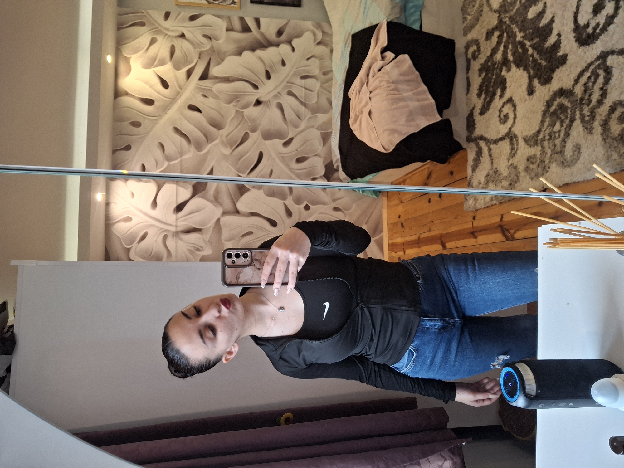 A woman is taking a selfie in front of a mirror. The mirror reflects a room with a bed and a patterned rug. The woman is wearing a black athletic jacket and blue jeans. In the foreground, there is a speaker and incense sticks on a white surface. The scene appears to be indoors. The location is unknown, and it is not possible to verify whether the scene is in Buhovo, Bulgaria.