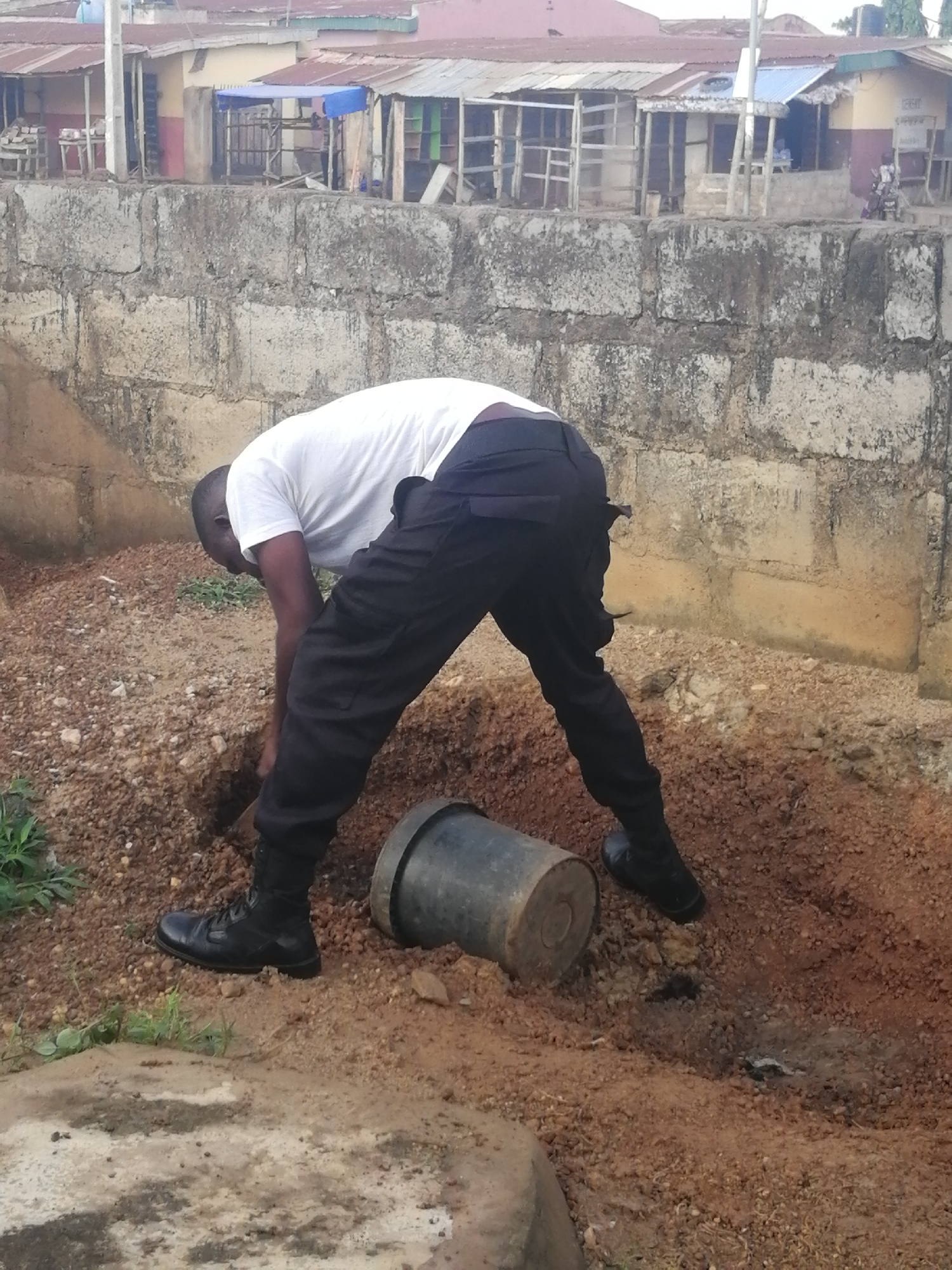 In Ajasse Ipo, Nigeria, a person dressed in a white t-shirt and black pants is digging a hole in the dirt. The individual is wearing black boots. A cylindrical object, possibly a pipe or container, lies on the ground next to the hole. The background features a low stone wall and some buildings with corrugated metal roofs. The ground is covered in loose soil and small stones.