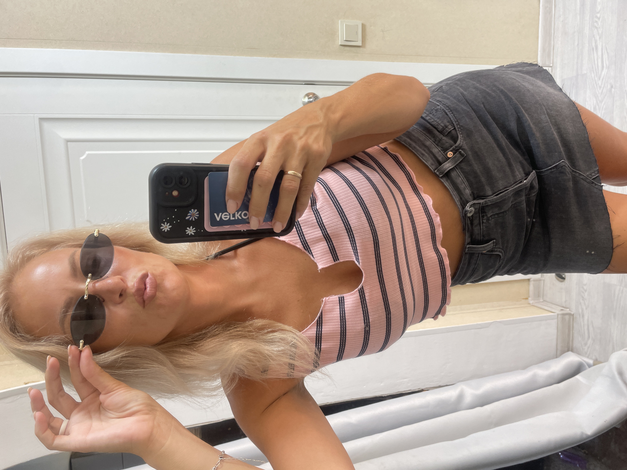In a selfie taken indoors, a blonde woman with fair skin is seen posing. She is wearing a pink and black striped crop top with a v-neck and a frayed hem, and a dark grey denim skirt. She is also wearing sunglasses and a gold ring on her left hand. Her right hand is holding her phone, which has a black case with a floral pattern and a blue card sticking out. The background appears to be a room with a white door and a light-colored wall. The lighting suggests it is daytime, possibly with natural light. There is no indication of the specific location within Baranovka, Russia.