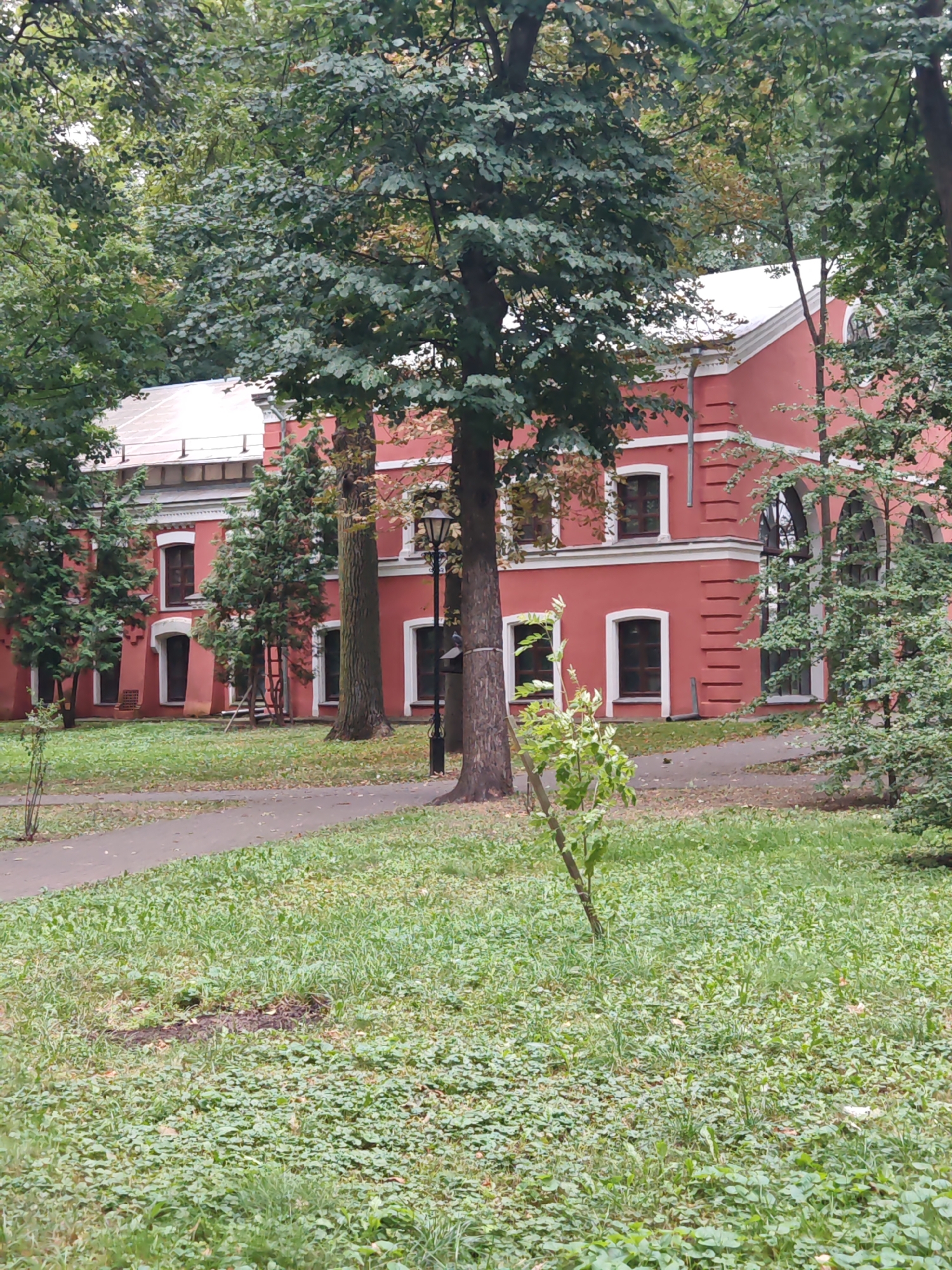 The image shows a portion of a large, two-story, red brick building with a white roof, situated in a park-like setting. The building has several windows with dark frames and white trim, and architectural details on its facade. A paved walkway runs in front of the building, bordered by a grassy area with scattered trees and foliage. A lamppost stands near the building. The overall scene suggests an outdoor environment, possibly a historical or public space, within the city of Homyel, Belarus.