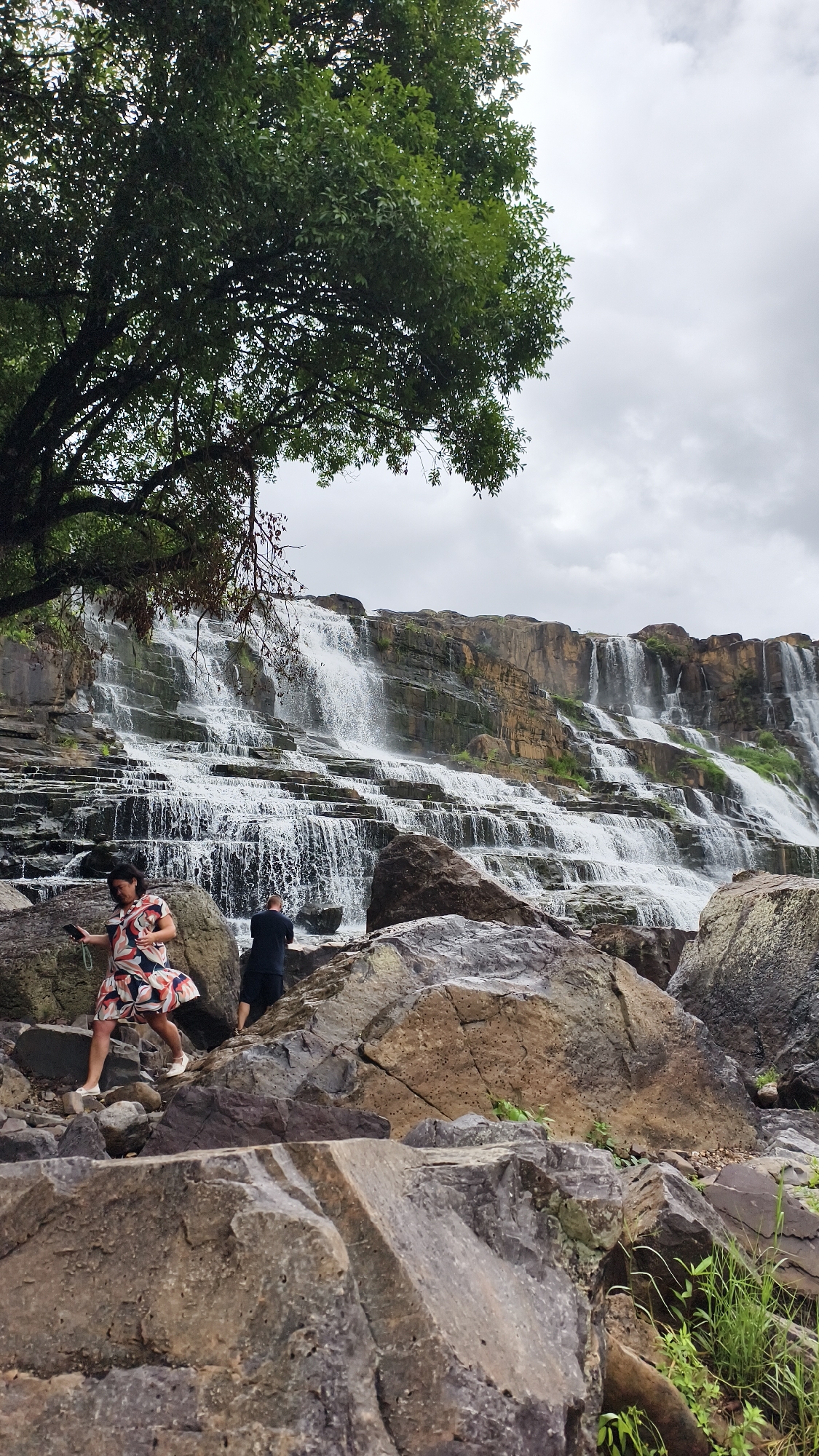 An image captures a tiered waterfall cascading down a rocky cliff face under an overcast sky. In the foreground, two individuals are positioned on a rocky outcrop. A woman in a vibrant, multi-colored dress is on the left, looking down at her phone while holding a green object. To her right and slightly further back, a person in a dark outfit with black shorts is facing away from the camera, also looking towards the waterfall. The area is characterized by large, textured rocks and patches of green vegetation. The waterfall itself is a prominent feature, with numerous streams of water flowing over ledges, creating a dynamic and impressive natural spectacle. The overall scene suggests a natural tourist attraction in a lush environment. This is a view of the Datanla Waterfall in the city of Da Lat, which is in the Lâm Đồng province, not Tân Phú.