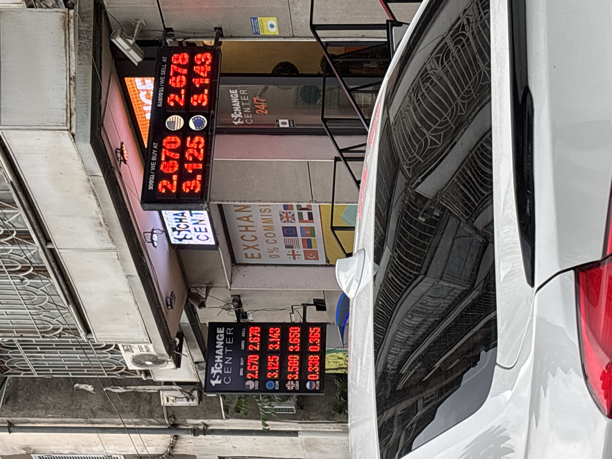 The image captures a street scene in Tbilisi, Georgia, dominated by an exchange center's currency rate displays and the rear of a white vehicle. The electronic signs show exchange rates for various currencies, including USD, EUR, GBR, and RUB. The signs are labeled with "BUY" and "SELL" rates in Georgian and English. The exchange center appears to be named "1st Exchange Center" and operates 24/7. Flags of various countries, including the EU, Turkey, and the US, are visible, suggesting the range of currencies dealt with. The street scene is characterized by architectural details and infrastructure, including buildings, wires, and air conditioning units. The white vehicle's rear is prominent, reflecting the surrounding buildings in its glossy surface.