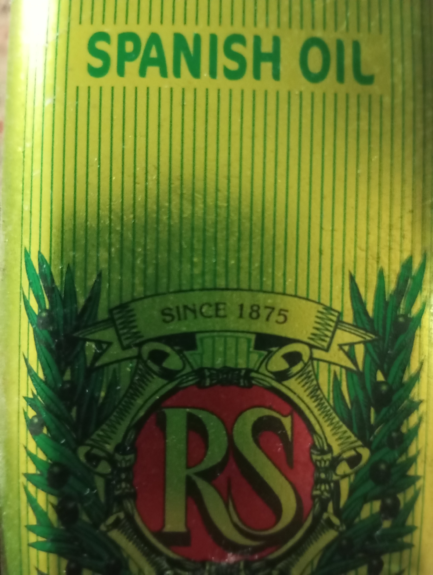 The image is a close-up of a product label. The label is bright yellow with thin green vertical stripes. At the top, in large green capital letters, it reads "SPANISH OIL". Below this, a green banner with a serrated edge reads "SINCE 1875". The banner is flanked by images of green olive branches with black olives. Centered below the banner is a circular emblem containing two large green letters, "RS", inside a red circle. The overall appearance suggests a vintage or traditional product. There is no indication of any specific location in Mansehra, Pakistan, or any people in the image.