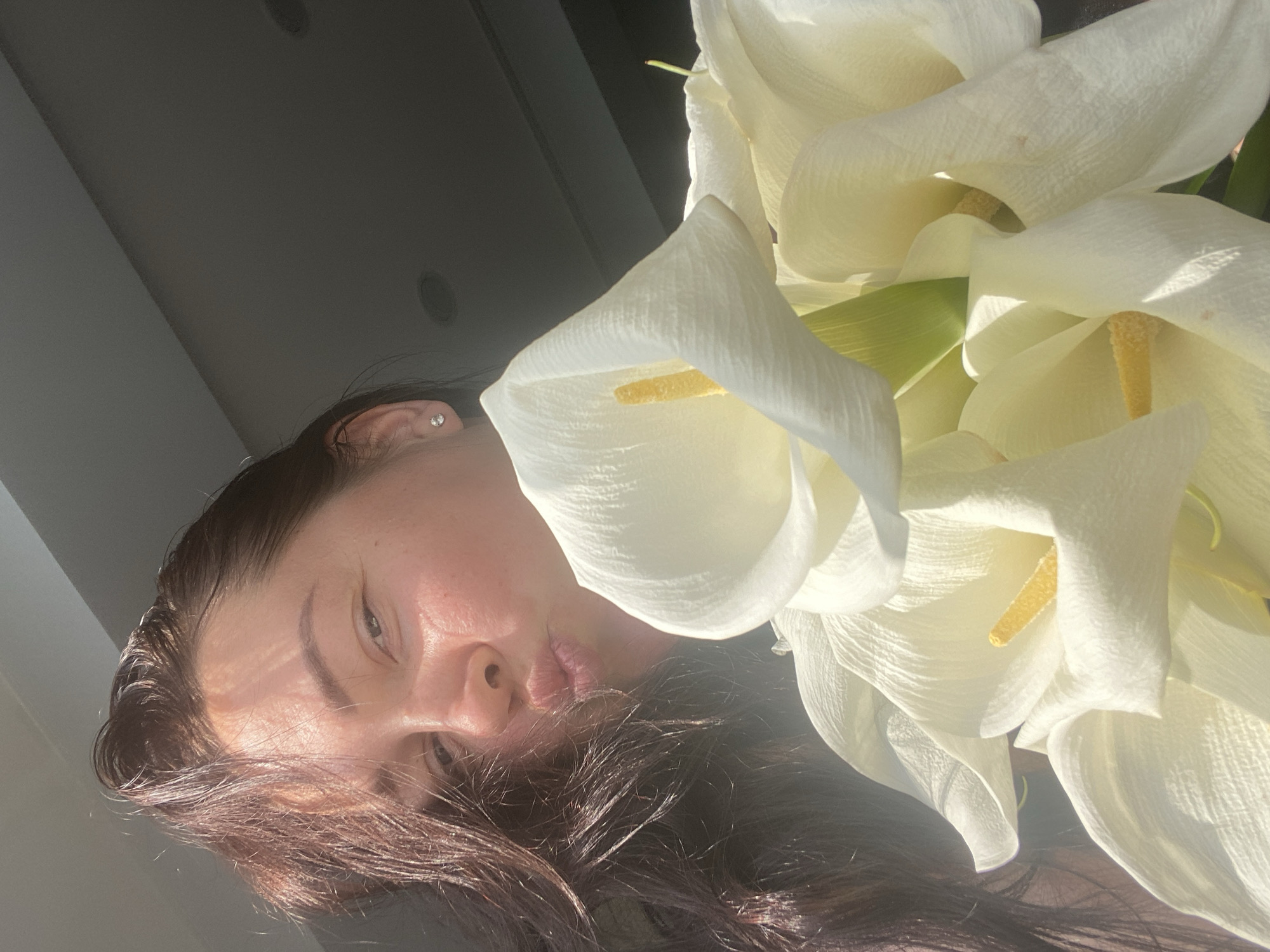 A woman with dark hair is holding a bouquet of white calla lilies. She is facing the camera, making a kissing face, with sunlight hitting her face. The background is a dark, out-of-focus interior space with a ceiling that appears to have recessed lighting fixtures. The scene is indoors, and there is no visible indication of the city of Sochi, Russia.