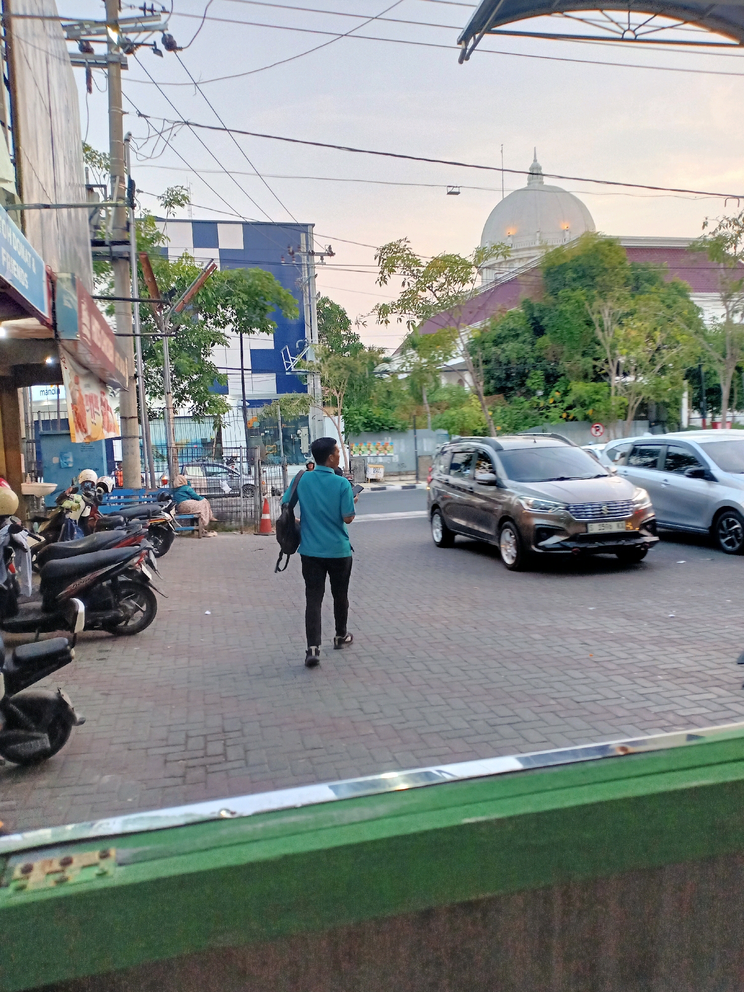 The image captures a street scene in Bojonegoro, Indonesia, likely during the late afternoon or early evening, given the soft lighting. A man wearing a teal shirt and black pants walks away from the camera, carrying a black backpack over his left shoulder and a small orange traffic cone in his right hand. He is walking on a brick-paved sidewalk adjacent to a street where a dark gray multi-purpose vehicle is driving. A light-colored vehicle is visible behind it.
To the left of the sidewalk, several parked motorcycles are lined up. Further down the sidewalk, a woman in a headscarf and light-colored patterned pants sits on a blue bench outside a building with a blue and white checkered facade.
In the background, a large white domed building with a pointed finial is visible, suggesting a significant public or religious structure. Trees with green foliage are interspersed throughout the scene, and overhead, a complex network of electrical wires crisscrosses the sky, supported by utility poles. The overall impression is one of a moderately busy urban environment with a blend of commercial buildings, infrastructure, and residential elements.