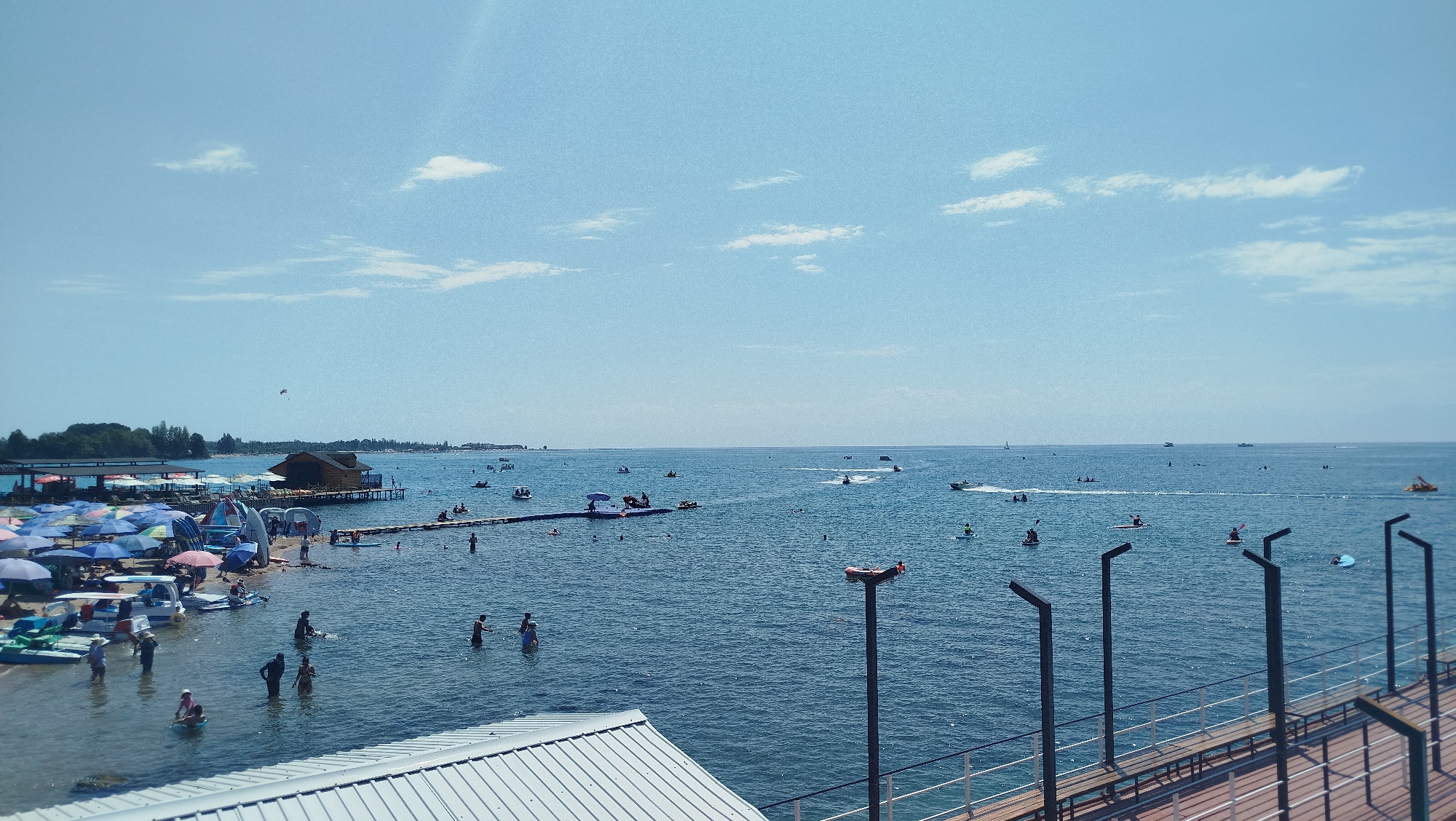 The image depicts a wide shot of a bustling beach scene on a clear day. The foreground features the roof of a building and a pier with dark metal posts and railings. The main focus is the blue ocean filled with various watercraft and people.
Numerous boats, including speedboats and what appear to be paddle boats, are scattered across the water. Several individuals are engaged in water activities, such as swimming and kayaking. A small wooden structure on stilts is visible in the middle ground, adjacent to a pier where people are standing.
The shoreline is lined with many colorful umbrellas providing shade, and various seating arrangements. People are seen wading in the shallow water near the shore and relaxing under the umbrellas. The sky is bright blue with a few scattered white clouds, suggesting fair weather. The overall impression is a lively recreational area on a sunny day.