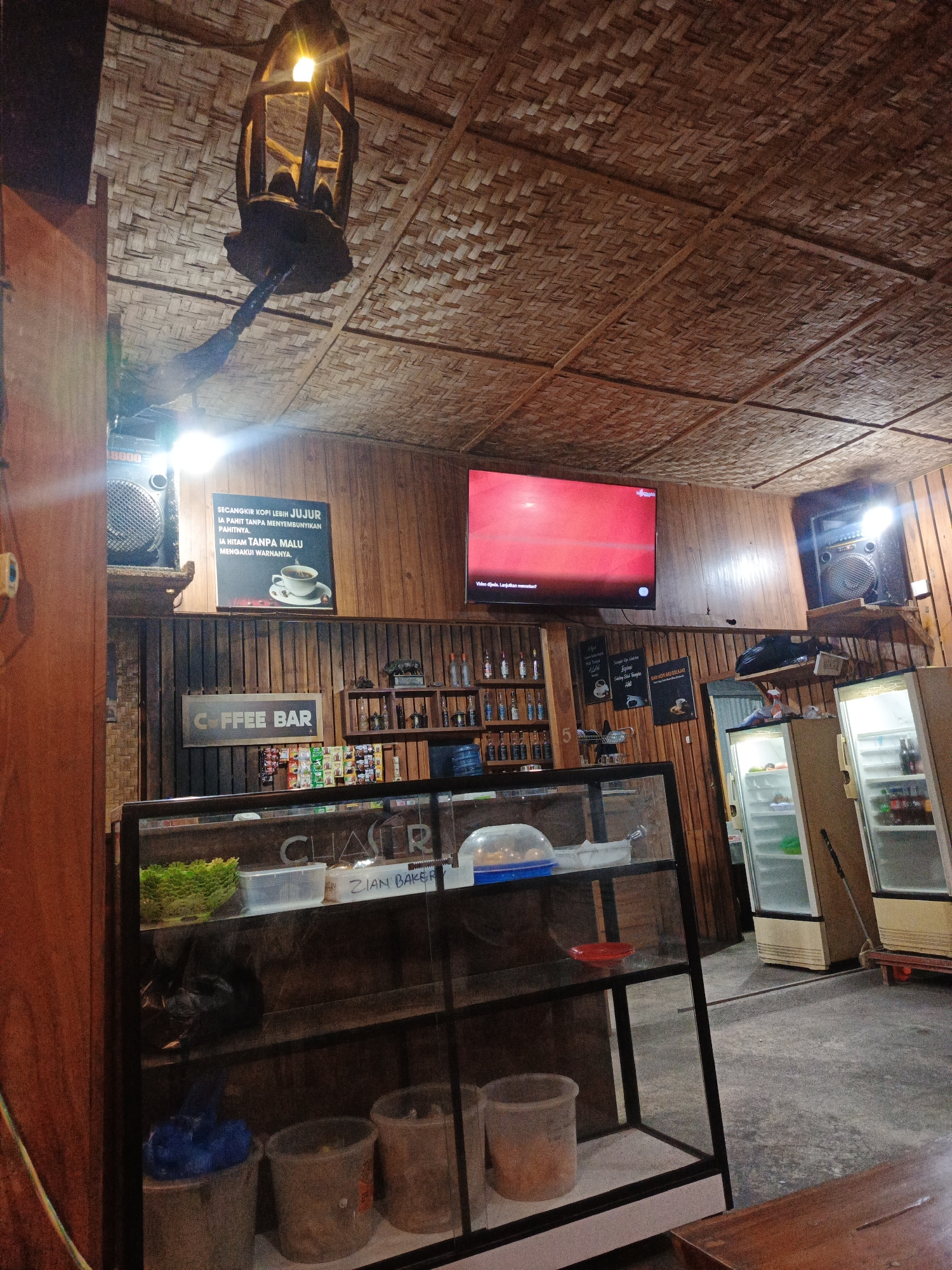 The image shows the interior of a coffee bar, possibly in Geulumpangmeujim-Jim, Indonesia. The room has wooden paneling on the walls and a woven ceiling. A decorative wooden light fixture hangs from the ceiling. A television screen is mounted on the wall, showing a paused video. Speakers are placed on either side of the screen.
A coffee bar sign hangs over a counter. A glass display case is in the foreground containing various items. There are refrigerators lined up towards the back of the room. A sign with coffee-related text is mounted above the counter.