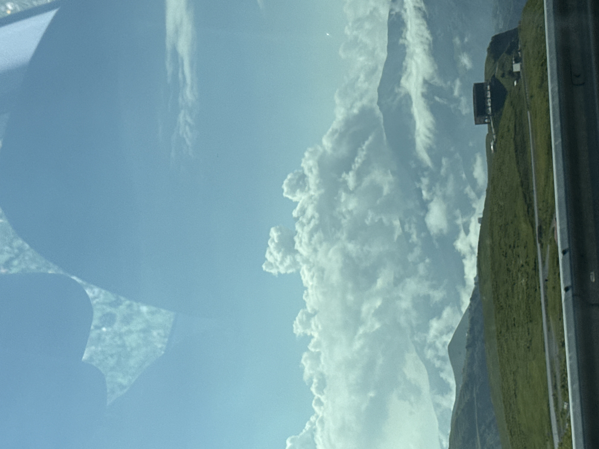 The image depicts a vast mountainous landscape under a partly cloudy sky. In the foreground, a metal guardrail runs horizontally across the bottom of the frame. Behind it, a winding road cuts through rolling green hills. Further in the distance, a dark, box-like structure with arched openings is situated on a hilltop. The middle ground is dominated by dramatic, voluminous white clouds that partially obscure the majestic mountain ranges behind them. The sky above is a pale blue, with some lighter cloud formations scattered near the horizon. The scene is likely captured from a moving vehicle, given the slight blur and the perspective. This appears to be the Gudauri Friendship Monument in Gudauri, Georgia, a popular tourist destination known for its panoramic views of the Caucasus Mountains.