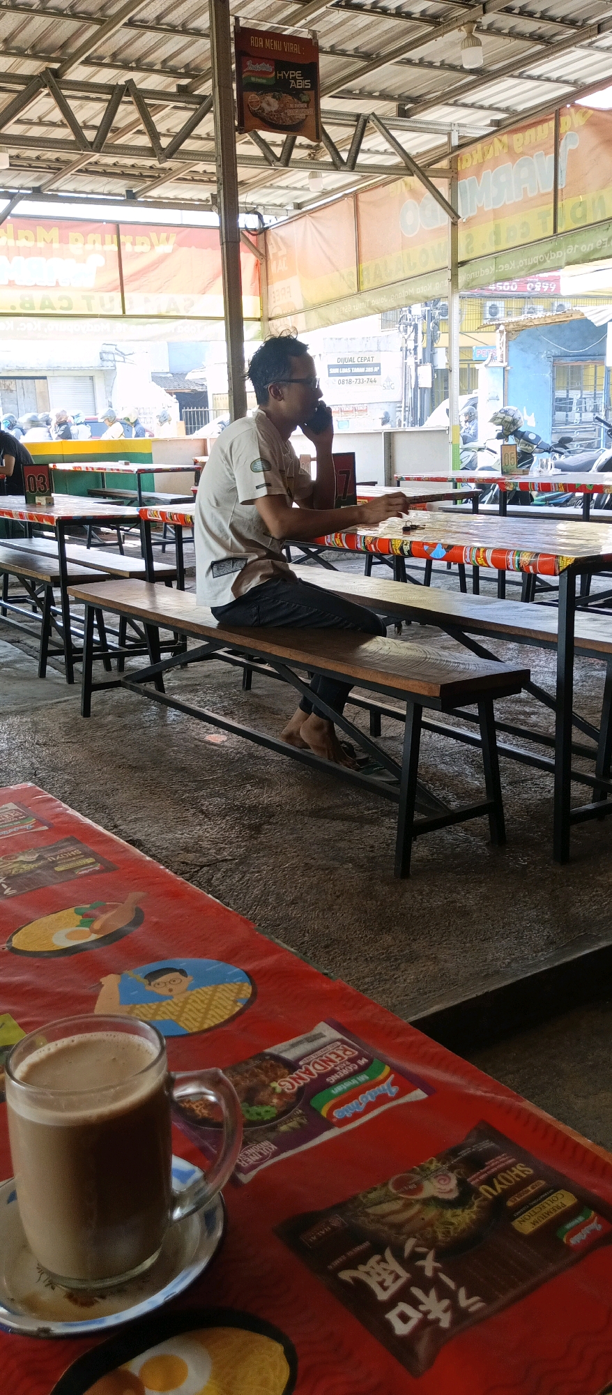 In a street-side food stall in Malang, Indonesia, a man sits on a wooden bench at a long table, holding a mobile phone to his ear. He is wearing a light-colored t-shirt and dark pants. The table is covered with a red tablecloth featuring cartoonish images and the Indomie logo. A glass of coffee and several packets of instant noodles are placed on the table. The stall appears to be open-air, with a metal roof supported by posts. In the background, motorcycles and other vehicles are visible on a street, suggesting an urban setting. The floor is concrete, and the overall atmosphere suggests a casual dining environment.