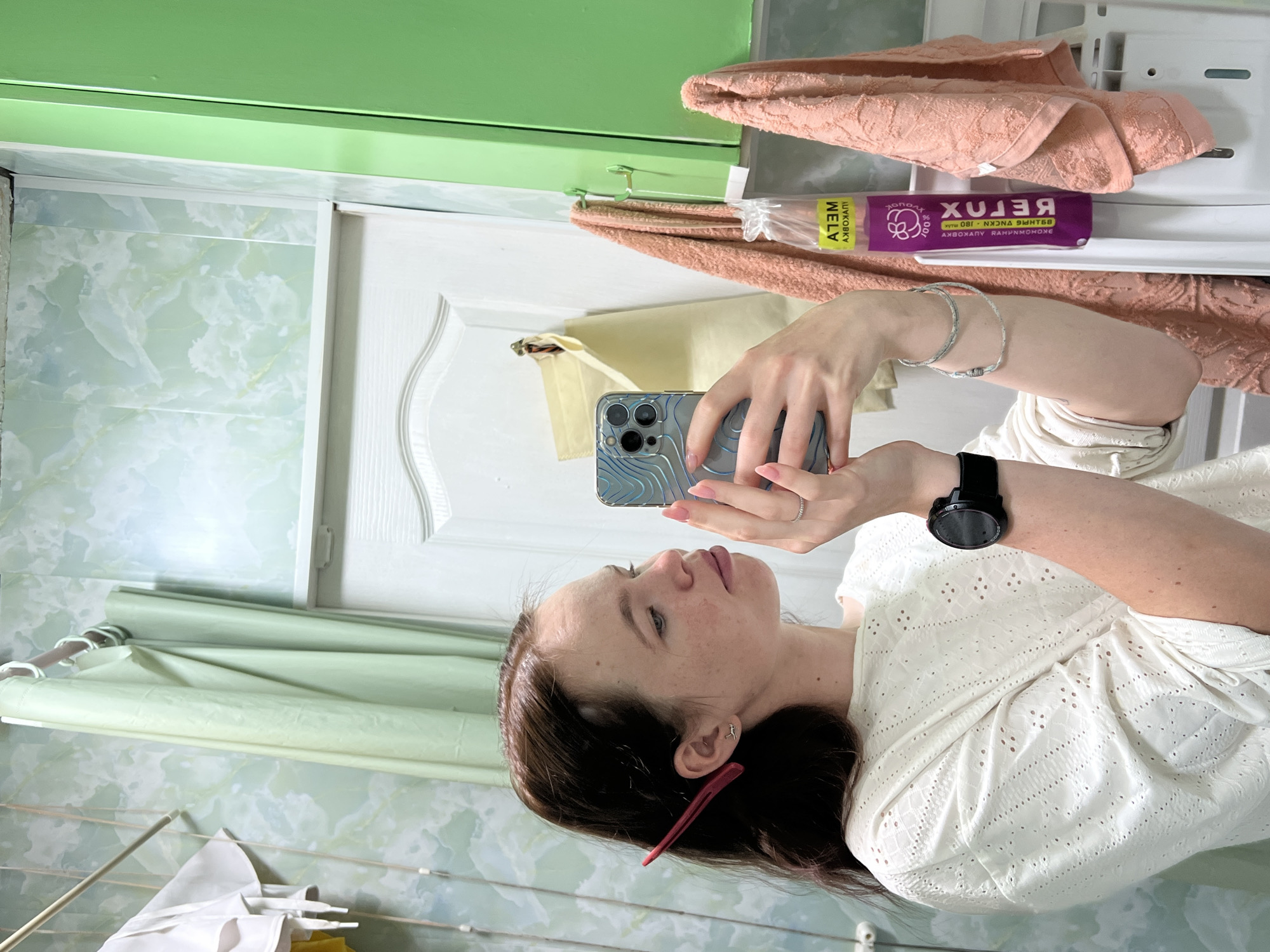 A fair-skinned woman with brown hair is taking a selfie in what appears to be a bathroom. She is holding a phone with a patterned blue case. She is wearing a white, eyelet-patterned top, a black smartwatch, and bracelets. A pink hair clip is visible in her hair. The background includes a green-painted wall and a white door, with pink towels and a purple box labeled "RELUX" on a shelf above. There is a green shower curtain, and what appears to be a laundry rack in the lower-left corner. The location is likely in Chaikovsky, Russia.