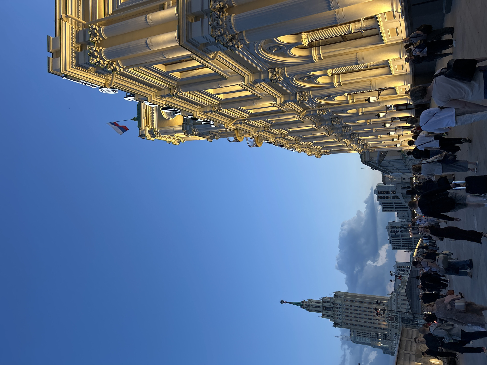 An evening urban scene depicts a bustling street or plaza, viewed from a low angle looking upwards. On the left, a large, ornate building with classical architecture, including columns, pilasters, and decorative cornices, is brightly illuminated by warm lights. A small flag, red, white, and blue, is visible hanging from this building. The sky above is a deep blue, transitioning to a lighter hue with some scattered clouds in the distance towards the right.
Numerous people are seen walking and standing on the ground level, appearing as pedestrians going about their evening. In the far background on the right, a distinctive tall skyscraper with a spired top, characteristic of Stalinist architecture, stands out against the sky. Other urban buildings are also visible further down the street.
The time of day is dusk or early evening, indicated by the artificial lights being on and the remaining daylight in the sky. The weather appears clear and pleasant. Part of an illuminated sign is visible on the ornate building, with characters forming "МОСКОВСКИЙ УНИВЕРСАЛЬНЫЙ МАГАЗИН" (Moscow Universal Store or GUM) being discernable, strongly suggesting the location is Moscow, Russia, specifically near GUM.