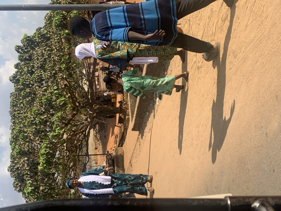This image captures an outdoor scene in Egbedi, Nigeria, under a bright sky with scattered clouds. The ground is an unpaved, light brown dirt surface, with visible shadows indicating strong daylight.
Four primary individuals are visible. In the mid-ground left, an adult female stands facing the camera. She wears a teal-patterned long dress with a matching headwrap and a white scarf draped over her shoulders. Her footwear consists of light-colored open-toed sandals. In the mid-ground center, a young girl walks away from the camera, wearing a long green dress patterned with white concentric spirals. Her hair is styled in braids. Behind her, an adult female walks in the same direction, wearing a colorful patterned long dress, a white hijab, and a white scarf. To the right of this adult female, a young boy walks in the same direction, wearing a blue and black horizontally striped long-sleeved shirt and dark trousers with dark clogs or sandals. All three walking individuals cast distinct shadows on the ground ahead of them.
In the background, a large, mature tree with dense green foliage dominates the center, providing shade. Beneath the tree and further back, indistinct figures and rudimentary structures, possibly market stalls or dwellings constructed from light-colored materials, are visible. A dark utility pole stands vertically on the right side of the frame. Concrete curbs or low barriers line parts of the path. The immediate foreground on the left shows a dark, vertical metallic structure, suggesting the photograph was taken from inside a vehicle or through a partial obstruction.