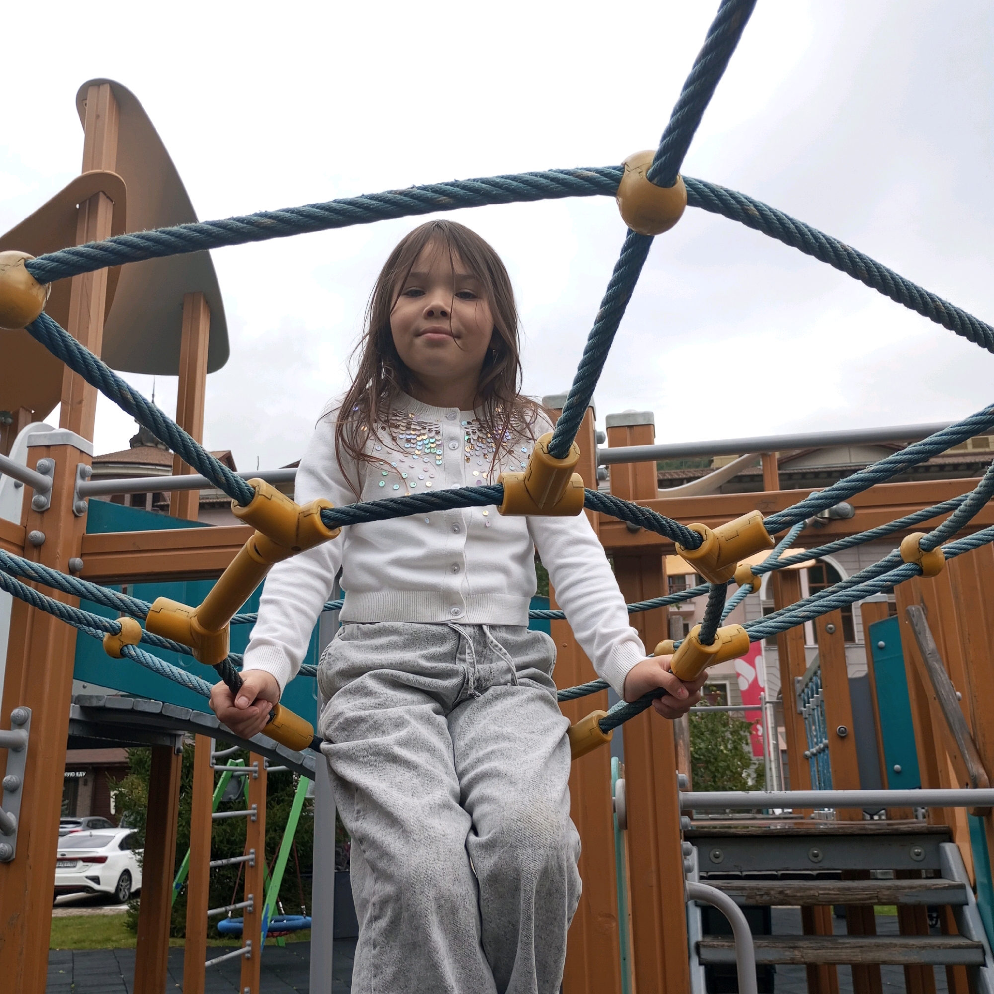 A young girl with long, dark hair is climbing on a rope net structure at a playground. She is wearing a white cardigan with sequins and grey sweatpants. The playground is made of wood and rope, with yellow connectors on the ropes. In the background, there are buildings and trees visible, suggesting an urban or suburban environment. The sky is overcast. The location is identified as Estosadok, Russia.