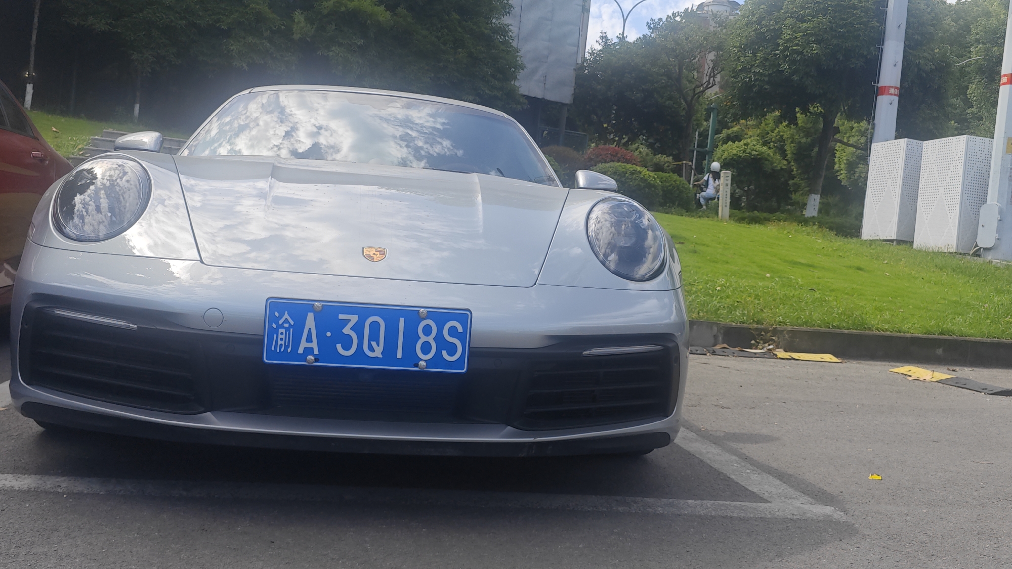The image shows a silver Porsche parked in a parking spot in Jiulongpo District, China. The license plate reads "渝A·3QI8S". A portion of a red car is visible on the left edge of the frame. In the background, there is a grassy area with trees and some buildings. A person wearing a white helmet can be seen sitting on a slope in the background. To the right of the person, there are two large white perforated metal structures. The weather appears to be overcast. There are yellow speed bumps in the lower right corner of the image.