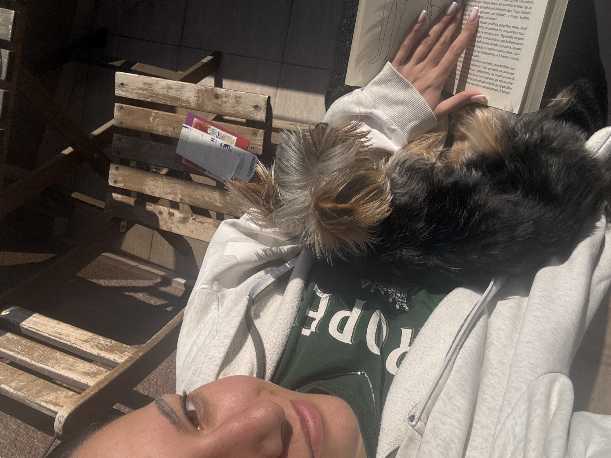 A woman is lying on her back outdoors, possibly on a lounge chair or similar seating arrangement. A small dog with brown and black fur is lying on her chest. She is holding an open book in her hand, with text visible in a language other than English. She is wearing a light grey hoodie over a dark green shirt with the word "ROPE" printed on it. A wooden chair or table is visible to the left of the frame, with a small object or package on it. The scene is brightly lit, suggesting it is daytime. The text in the book is in the Cyrillic alphabet, and the word "СЛИКОН" is printed on the package on the chair. I am unable to determine the exact location in the city of Varna, Bulgaria.