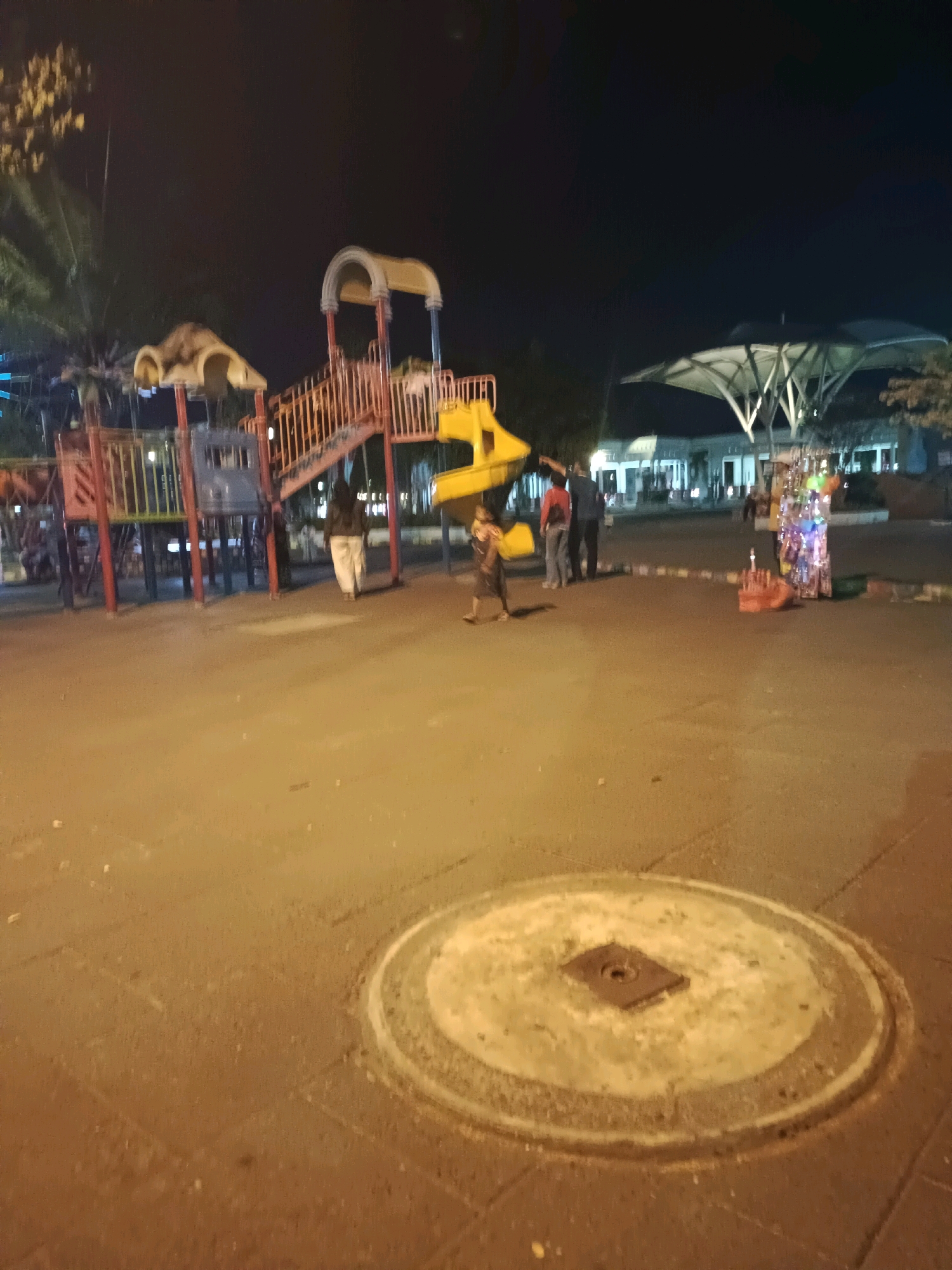 The image depicts an outdoor playground scene at night, possibly located in Lamongan, Indonesia. In the foreground, a circular concrete structure with a metal plate is visible on the asphalt ground. To the left, a colorful playground structure with slides, stairs, and platforms is present. Several people are scattered around the playground area. A person in light-colored pants and a dark top walks away from the playground towards the right. Another individual, a child, is seen running in the middle ground. Further to the right, three adults stand together, with one pointing. In the background, a building with an arched, canopy-like roof is illuminated. A stall with colorful lights and products is set up near the adults. The overall lighting suggests it is nighttime, with artificial lights illuminating the scene.