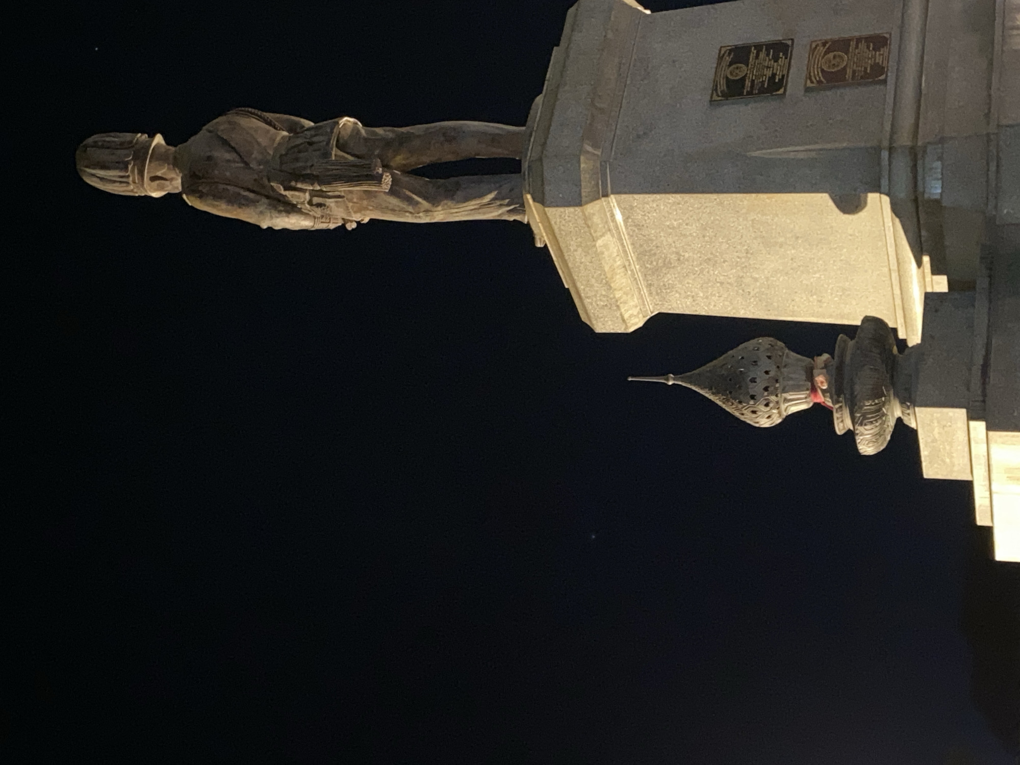 At night in Baan Don Sai, Thailand, a bronze statue stands atop a stone plinth. The statue depicts a male figure in historical dress, wearing a hat and holding what appears to be a rolled document. Two plaques are affixed to the plinth above a decorative roof-like structure. The night sky is dark.