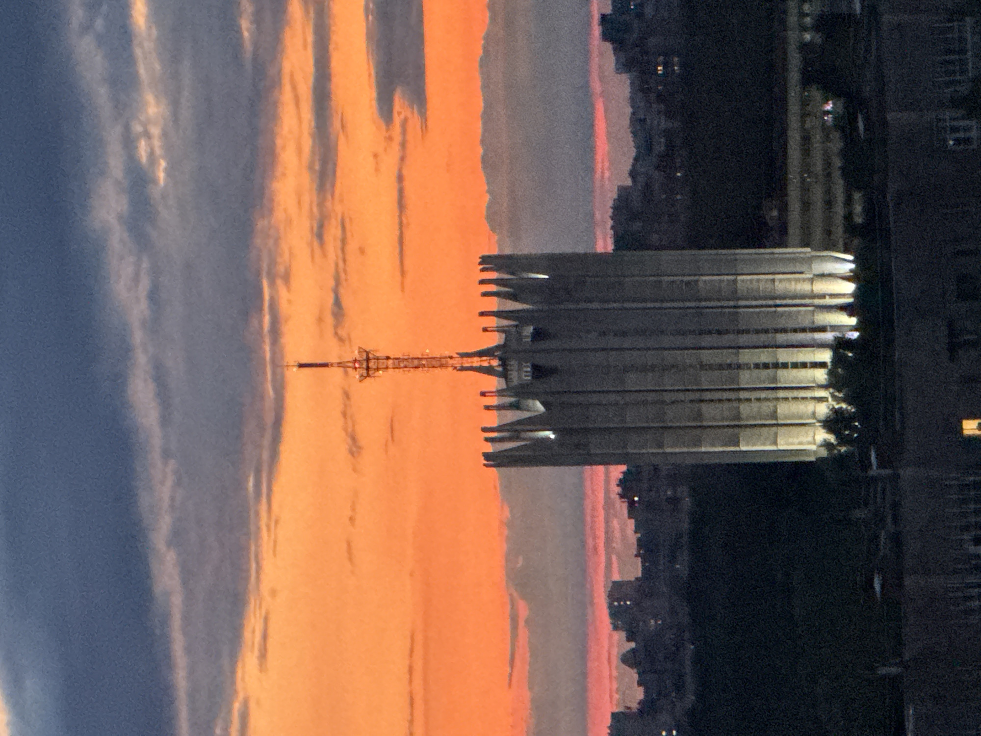 A telephoto shot captures a tall, angular building topped with a radio tower against a vibrant sunset sky. The building is constructed of concrete and features a distinctive crown-like, serrated roofline. Lights illuminate the lower sections of the structure, casting a warm glow. In the background, a dense cityscape is visible, with buildings silhouetted against the twilight. A strip of dark trees lines the foreground, obscuring the lower portions of the city and the base of the main building. The sky is a dramatic gradient of fiery orange near the horizon, transitioning to deep blue and gray at the top, with streaks of clouds contributing to the atmospheric effect. The overall impression is one of urban architecture bathed in the dramatic light of dusk.