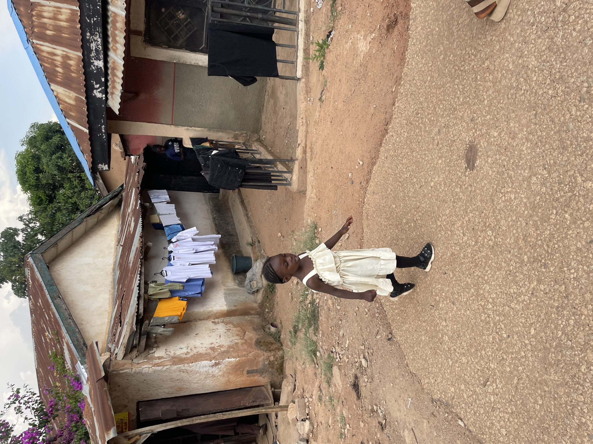 A young girl in a white dress stands on a paved road in what appears to be Jos, Nigeria, as the scene features a dilapidated building structure with a corrugated metal roof, clothing hung out to dry, and the environment reflects the area's characteristic reddish-brown soil. The photograph shows what is common in low-income neighborhoods of the city. The little girl in the white dress stands out from the backdrop.