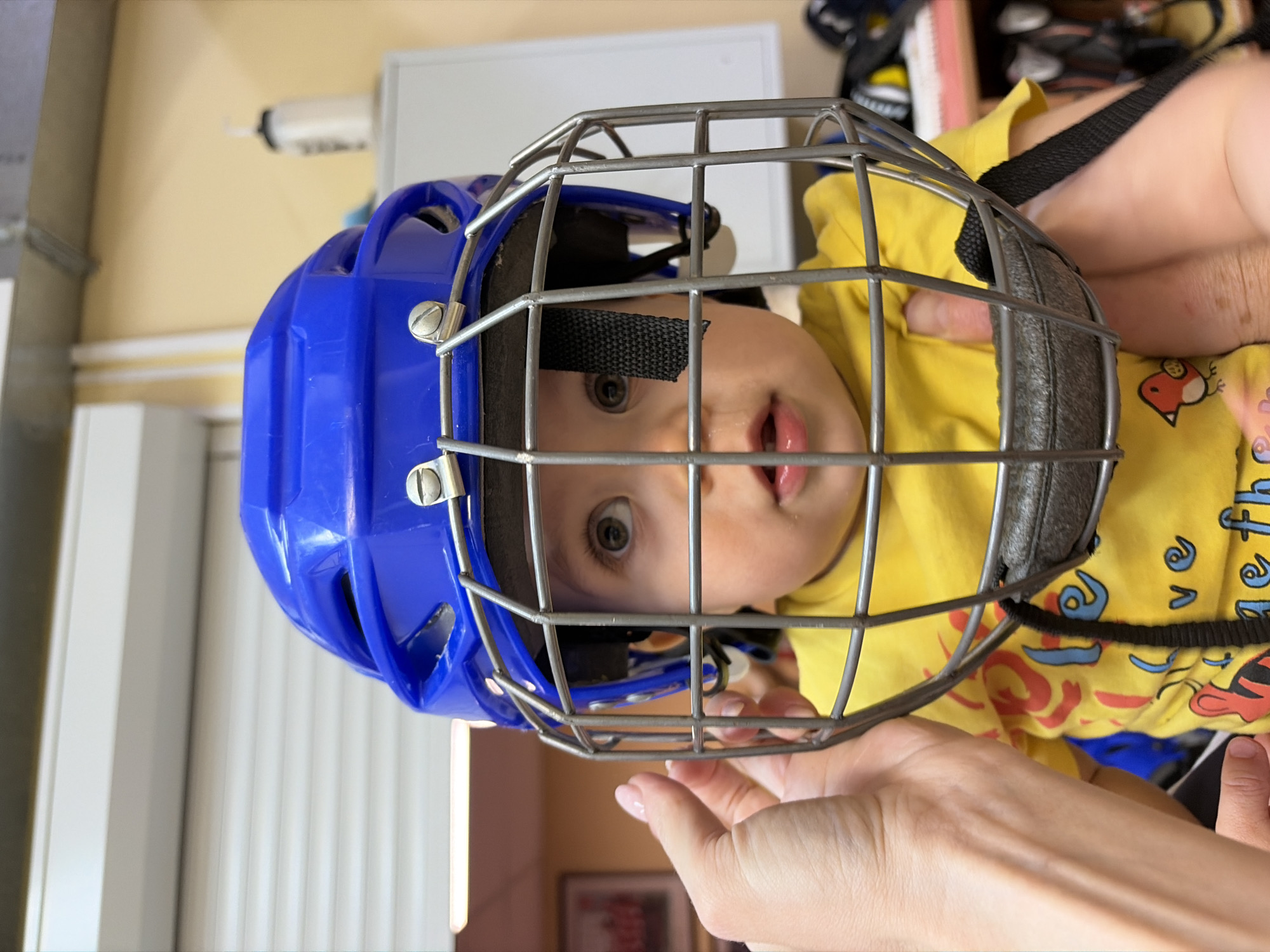 A young child wearing a blue and gray hockey helmet is being held by an adult. The child has light skin, light hair, and is wearing a yellow shirt with cartoon designs. The adult's hands are visible, indicating they are holding the child. The background includes a light-colored wall and a partially visible window with blinds. The location is Chaikovsky, Russia.