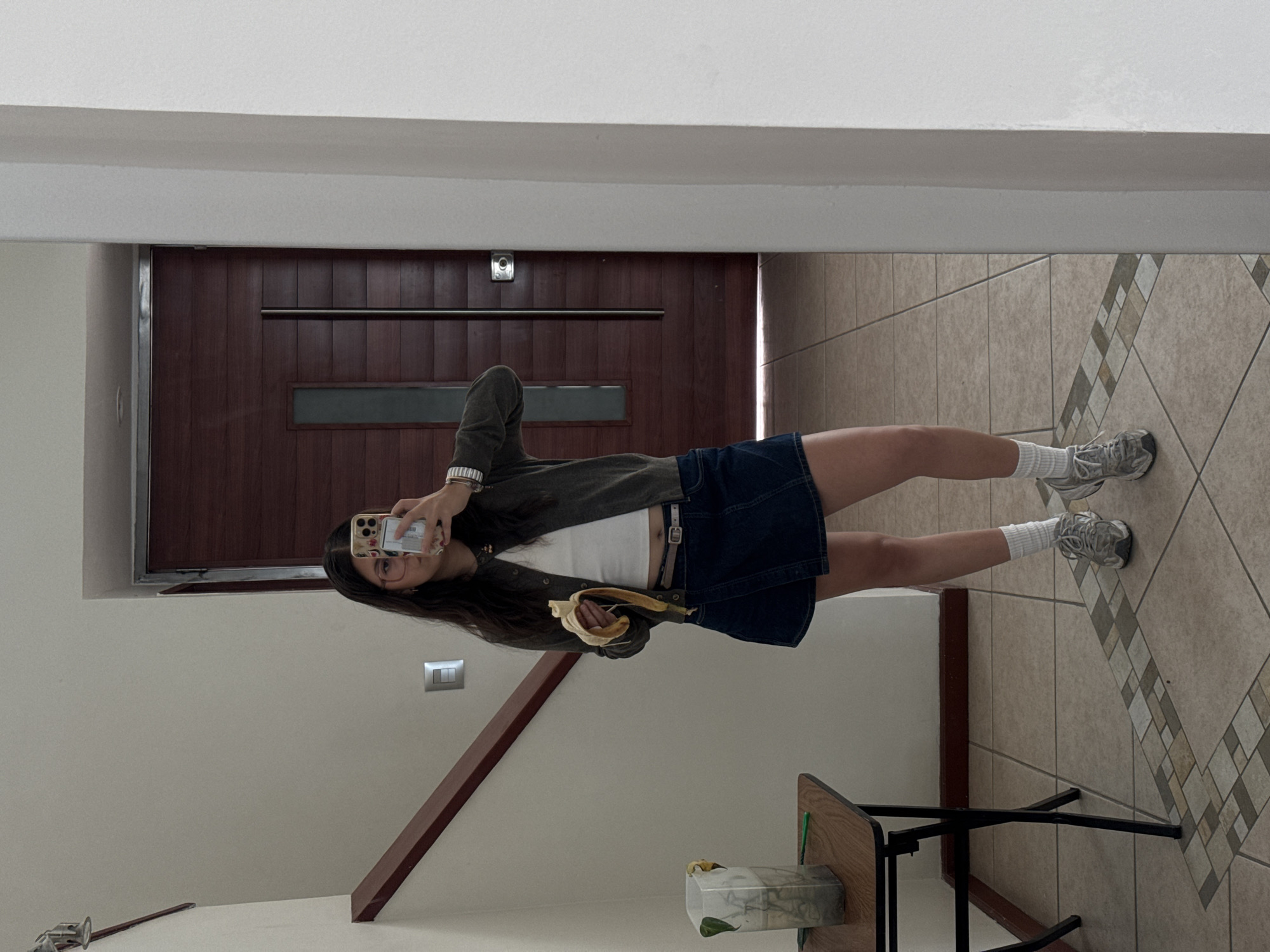 An upright, eye-level indoor shot shows a young woman taking a mirror selfie, holding a partially peeled banana in her left hand. She wears a dark olive green long-sleeved top or jacket over a white crop top, a dark denim mini-skirt, white socks, and grey athletic shoes. Her dark hair falls over her shoulders, and she wears glasses, with her face reflected in the smartphone she holds up. Her right wrist is adorned with a watch and bracelets.
The background reveals a hallway or entrance area of a home. Behind her is a tall, dark brown wooden door with a horizontal metal handle and a narrow, rectangular window. Above the door, a small, circular light fixture is visible on the wall. The floor is tiled with light beige squares, featuring a decorative strip of smaller, multi-toned mosaic tiles running diagonally across the scene. To the left, a light-colored wall with a light switch and a wooden handrail suggest a staircase or architectural feature. To the right, a small table or stool with a black metal frame and a wooden top holds a clear glass vase or container with a green plant inside. The overall lighting suggests it is daytime. The woman appears to be smiling slightly, conveying a casual and relaxed mood. No discernible text is visible on any objects or the phone screen. The location context provided is Heroica Puebla de Zaragoza, Mexico.