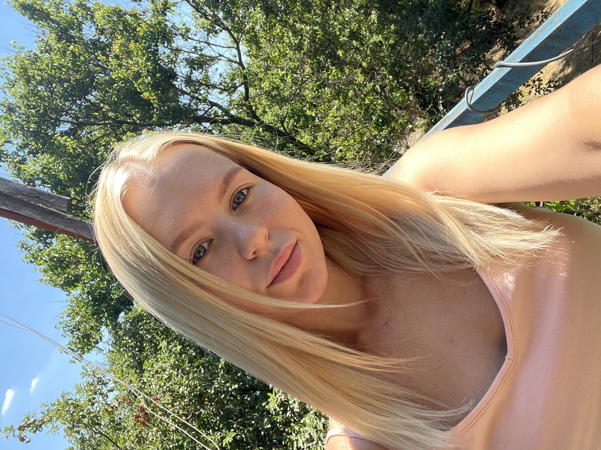 The media file is a selfie of a young woman with blonde hair. She is outdoors, with green trees and a blue sky visible in the background. The sun is shining, casting a warm light on her face and hair, suggesting it is daytime. She is wearing a pink top. Her expression is relaxed and pleasant. The scene appears to be a casual, outdoor setting, possibly a garden or park. There are no visible objects of note other than the natural surroundings and her clothing. No text is visible in the image. The image is taken from a slightly angled perspective, with the woman's face tilted towards the camera.