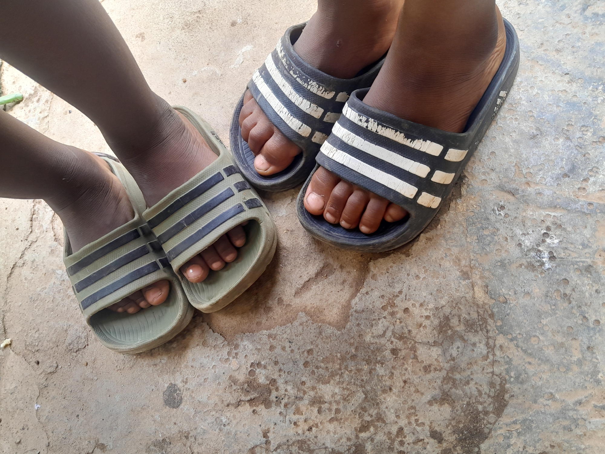 The image displays a close-up, overhead view of two pairs of feet wearing Adidas slides, resting on a textured, concrete ground. The ground appears weathered with some discoloration and embedded debris. One pair of slides is dark blue with white stripes, and the other is olive green with dark stripes. The feet are bare and appear to belong to children, judging by their size and the shape of their toes. The skin tone of the feet is dark. The overall lighting suggests it is daytime. The scene is likely outdoors in a casual setting, possibly a residential area or a public space with a concrete floor in Madalla, Nigeria.