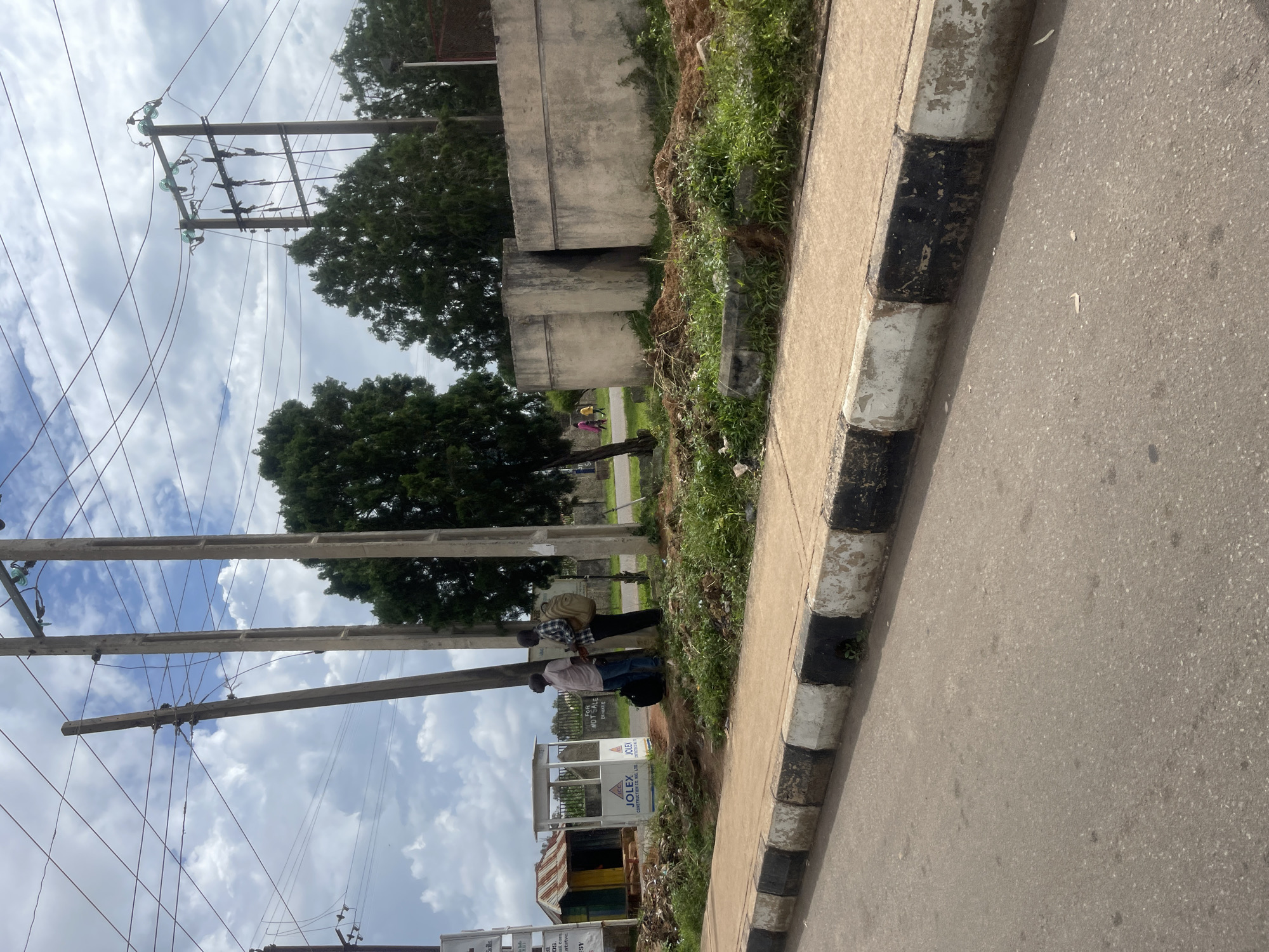 A street scene in Bar Jirgi Summa, Nigeria, shows two men standing on a sidewalk next to a small bus stop shelter and utility poles. The man on the left is wearing a light-colored shirt and jeans, with a dark bag at his feet. The man on the right is wearing a plaid shirt, dark pants, and a backpack. Behind them are a few concrete structures and trees. The sidewalk is bordered by a road with a painted curb. The sky is cloudy.