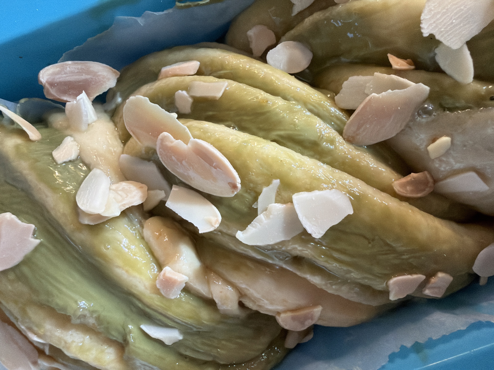 A close-up, high-angle shot displays a green-hued, glossy braided loaf or pastry, likely a dessert. The loaf is arranged in layers or strands, with what appear to be sliced almonds sprinkled on top. The background includes a light blue, possibly a tray or container. The image does not provide information about Bretton, United Kingdom or any people, actions, or interactions in that location.