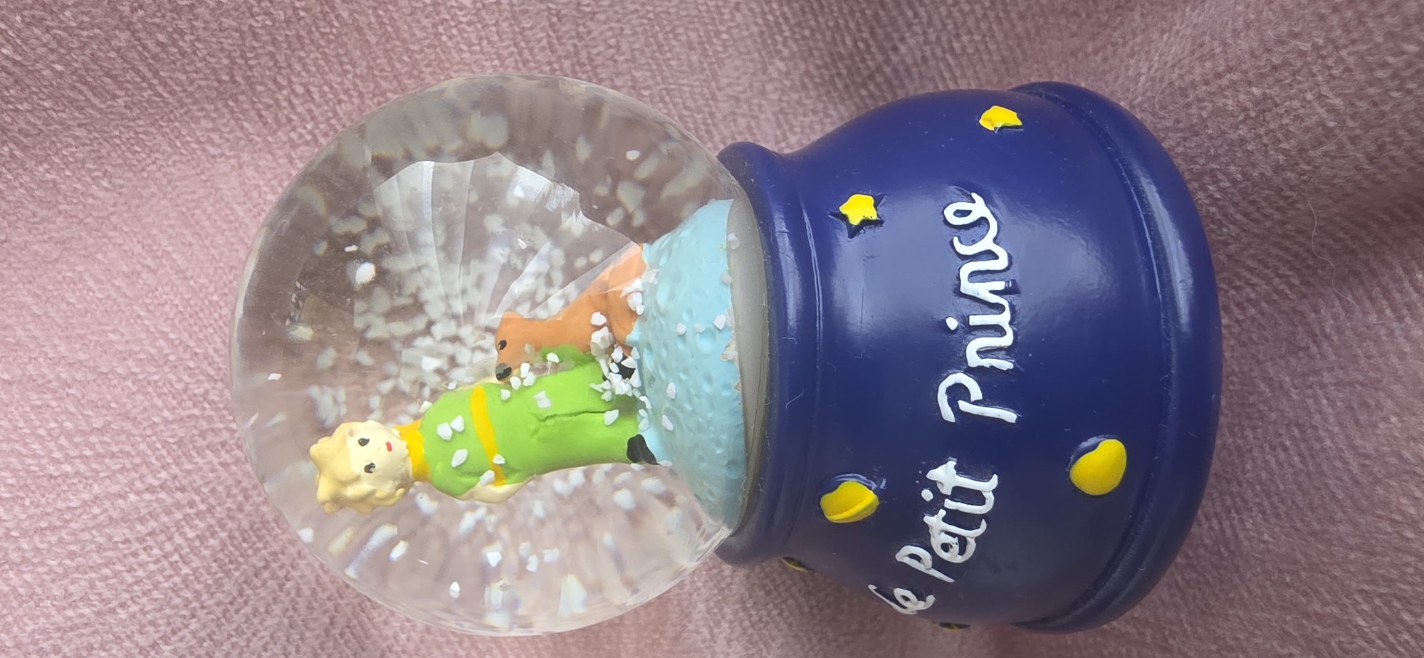 The image shows a close-up of a snow globe that features a figurine of the Little Prince standing with a fox on a blue, cloud-like base. The snow globe is filled with white glitter that appears to be falling. The base of the snow globe is dark blue and decorated with yellow stars and the words "Le Petit Prince" in white script. The snow globe is placed on a textured pink surface, likely a fabric. There is no information in the image to suggest a location in Sumgayit, Azerbaijan.
