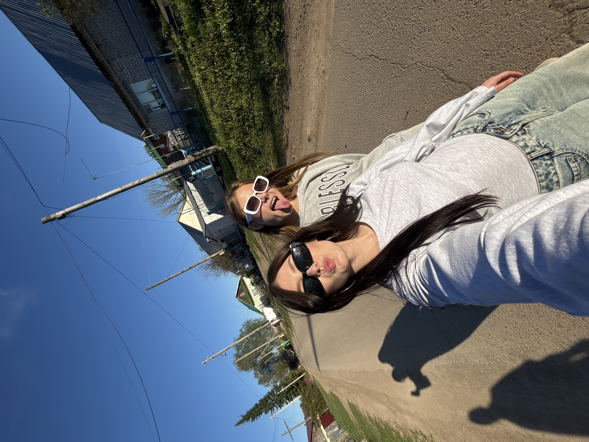 Two women are lying on a paved road. One woman wears sunglasses and sticks her tongue out. The other woman wears sunglasses and purses her lips. The women are wearing long-sleeved shirts and jeans. Houses and utility poles are visible in the background. The sky is clear and blue. The scene is set in Фоки, Russia.