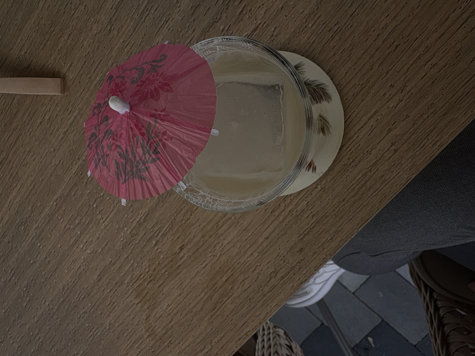 On a wooden table, a glass of light-colored liquid with a large ice cube sits on a coaster with a floral pattern. A small, red paper umbrella is inserted into the glass. Next to the glass, on the table, is a rectangular wooden object. Part of a gray cushion and wicker chair are visible in the lower right corner of the image. The photo was likely taken at an outdoor cafe or bar in Kragujevac, Serbia.