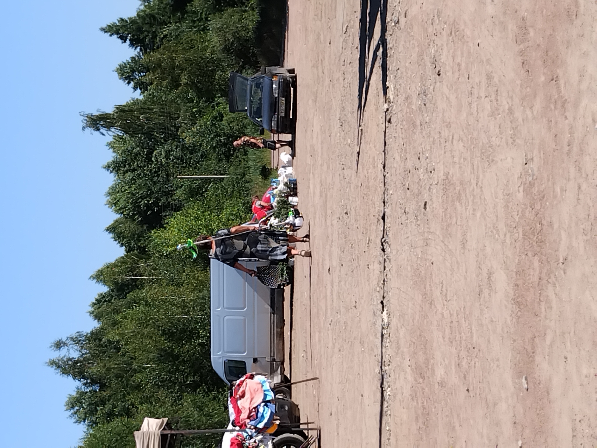 A person walks across a dusty lot on a sunny day, carrying a long object with a green attachment over their shoulder and a patterned bag in their other hand. They are dressed in a dark, patterned dress and sandals. Behind them, a light-colored van is parked, and to the right, a dark-colored car with its trunk open is visible. A pile of clothing is draped over a stand on the left side of the image, and another person stands near the car on the right. The background is filled with a dense line of green trees under a clear blue sky. This scene appears to be located in Irshansk, Ukraine.