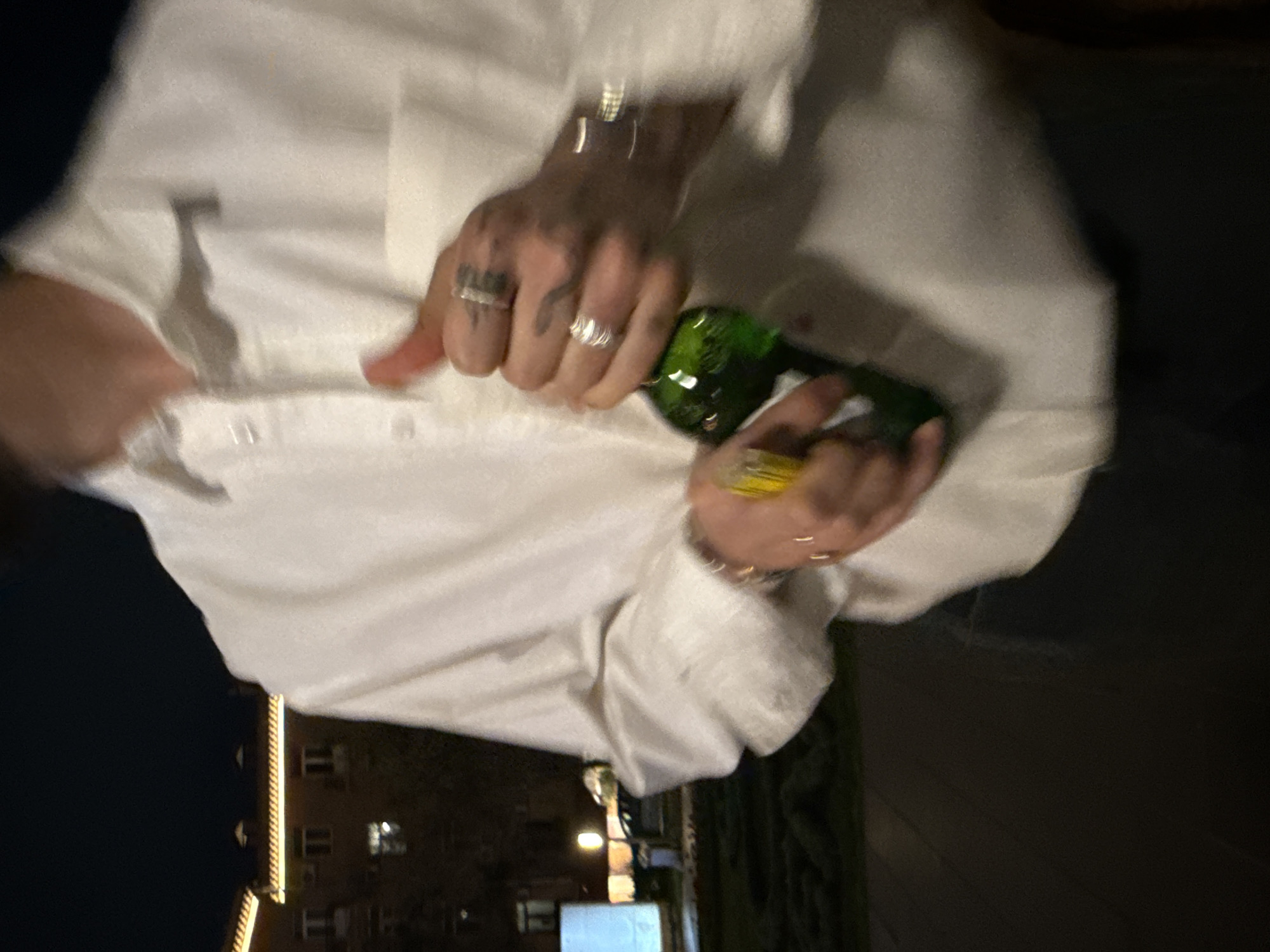 A person wearing a white, button-down shirt and multiple rings on their fingers is holding a green glass bottle with a yellow liquid inside. The scene is outdoors, possibly in the city of Yerevan, Armenia, suggested by the building with illuminated windows in the background. The overall lighting indicates it is nighttime. The person is holding the bottle with both hands, fingers curled around it.