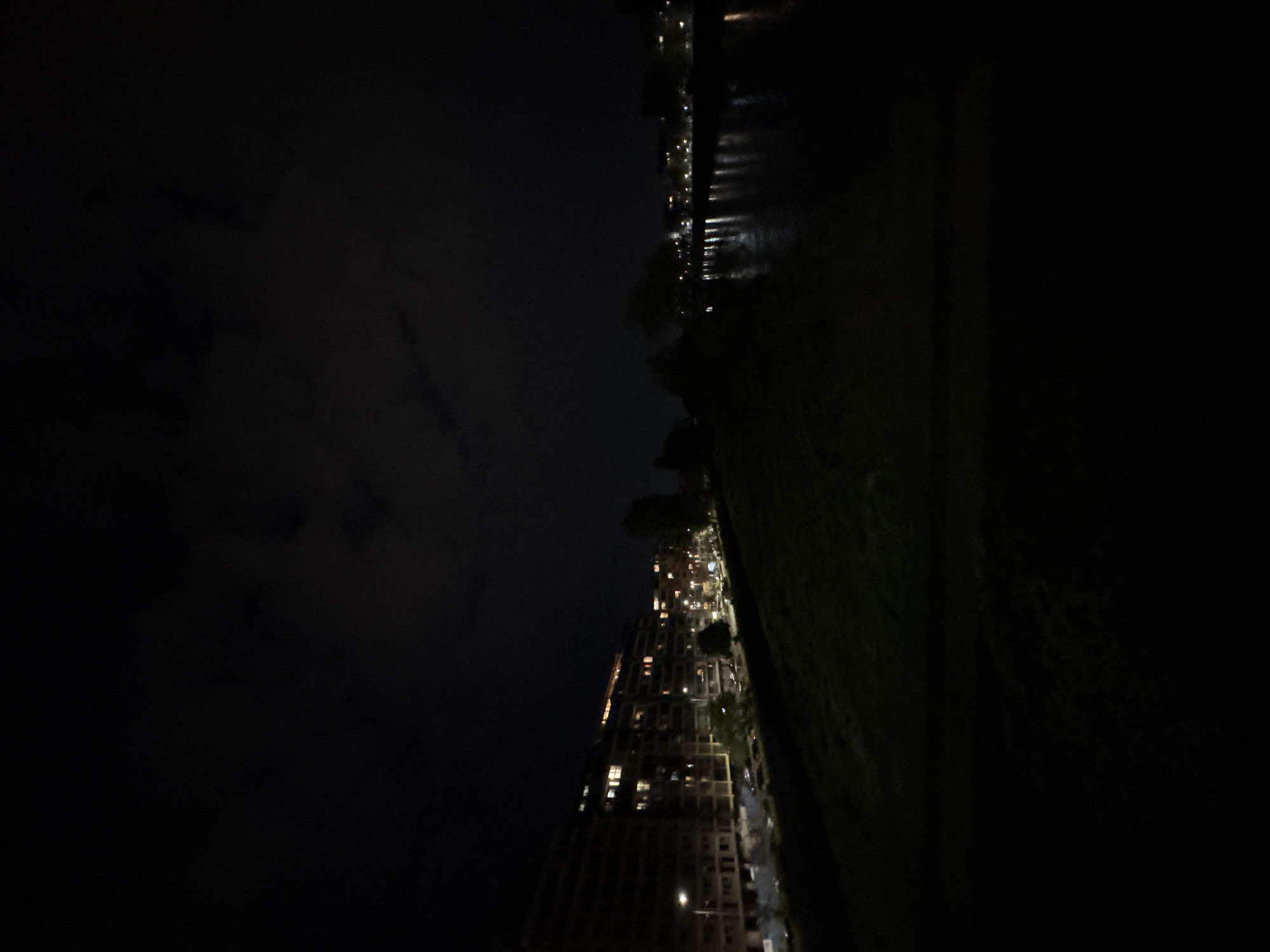 This vertical night-time image captures a cityscape viewed from a high vantage point, presenting a stark contrast between illuminated urban elements and pervasive darkness.
The scene, when viewed horizontally, shows a dark, cloudy sky on the left. In the middle, a dense strip of urban development runs horizontally across the frame. This area is brightly lit, showcasing numerous buildings with glowing windows, suggesting residential or commercial activity. Smaller, punctiform lights are scattered throughout, likely from streetlights or vehicles, delineating streets and structures. The architecture appears modern, with multiple stories visible.
On the right side of the frame, a dark body of water, likely a river, stretches along the illuminated urban edge. This water reflects the city lights, creating shimmering vertical streaks. Beyond the water, a dark, undefined landmass, possibly a park or natural area with trees, completes the composition.
There are no identifiable people or specific activities taking place, contributing to a sense of nighttime tranquility. The time of day is clearly night, evidenced by the darkness and reliance on artificial lighting. The weather appears to be overcast or partly cloudy, with subtle cloud formations discernible against the dark sky. Given the location context of Plovdiv, Bulgaria, this is likely a view of the city along the Maritsa River. No visible text is present. The overall mood is quiet and contemplative, highlighting urban life after dark.