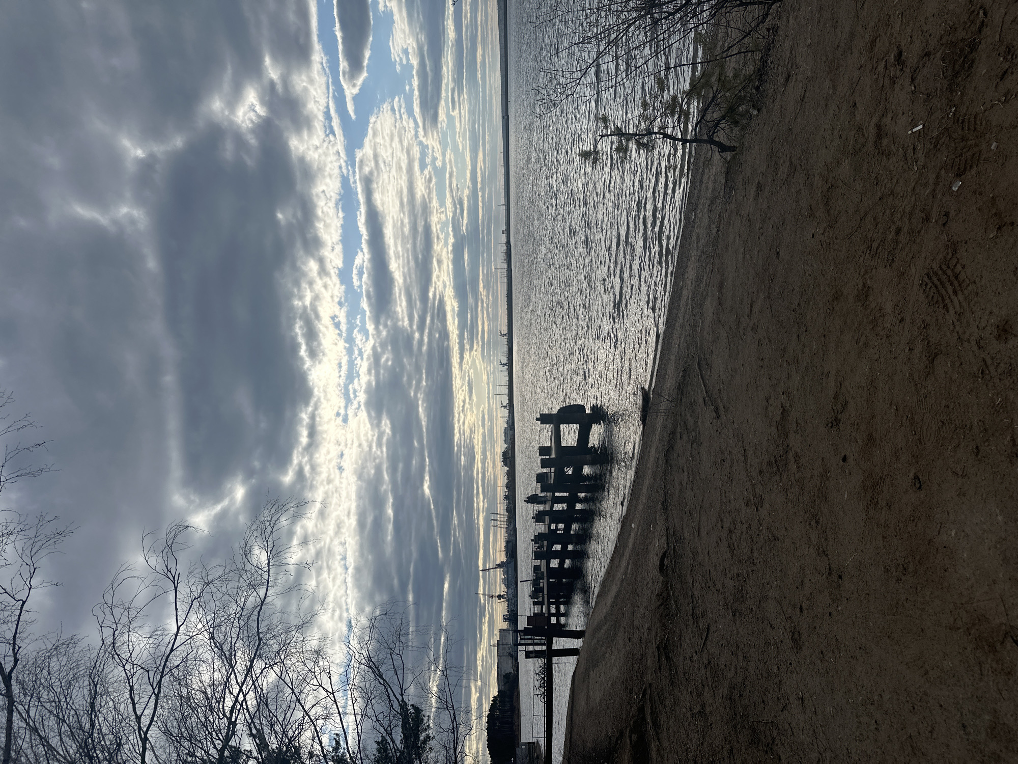 The image depicts a lakeside scene in Chaikovsky, Russia, under a sky with dramatic, layered clouds and patches of blue. The sun is low, casting a bright glow between the cloud layers. In the middle ground, the remains of a wooden pier extend into the water. The pier's pilings are weathered and decaying. The lake water is textured with small waves and reflects the light from the sky.
In the foreground, a sandy beach stretches along the right edge of the image. To the left, bare tree branches reach upwards, silhouetted against the sky. In the distant background, the skyline of Chaikovsky is faintly visible, with industrial structures like cranes and buildings. The general impression is one of a quiet, slightly bleak, natural environment juxtaposed with industrial presence.