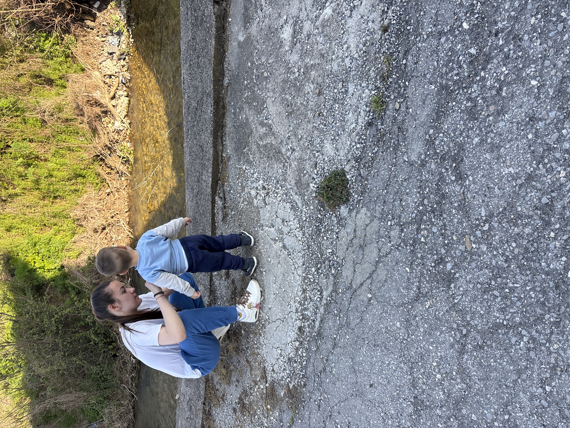 A woman and a young child are positioned on a concrete structure, likely a bridge or raised platform, adjacent to a shallow stream or river in Siva Reka, Bulgaria. The woman is in a crouching position, appearing to interact with the child. The child is standing upright, facing the woman. The stream flows alongside the concrete structure, and vegetation is visible along the stream bank. The concrete surface shows signs of wear and tear.