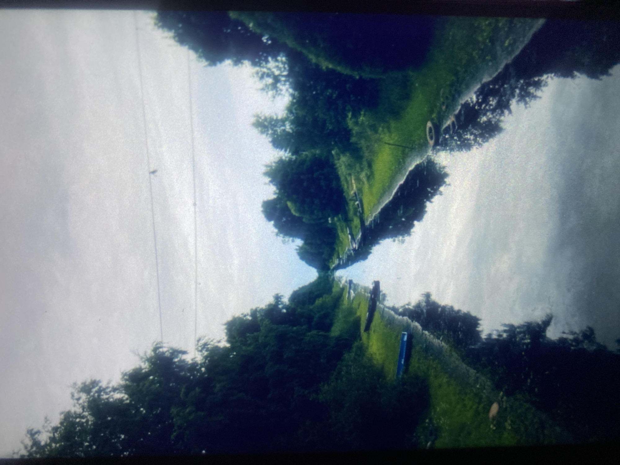 The image depicts a calm, narrow waterway flanked by lush green trees and vegetation on both sides. The water reflects the overcast sky and the surrounding foliage, creating a symmetrical, mirrored effect. Several small, dark-colored boats are visible pulled up on the grassy bank to the left of the waterway. In the distance, the waterway appears to converge with a hazy horizon. A power line is visible stretched across the sky. The scene appears to be a rural or semi-rural area, possibly a canal or a natural waterway.