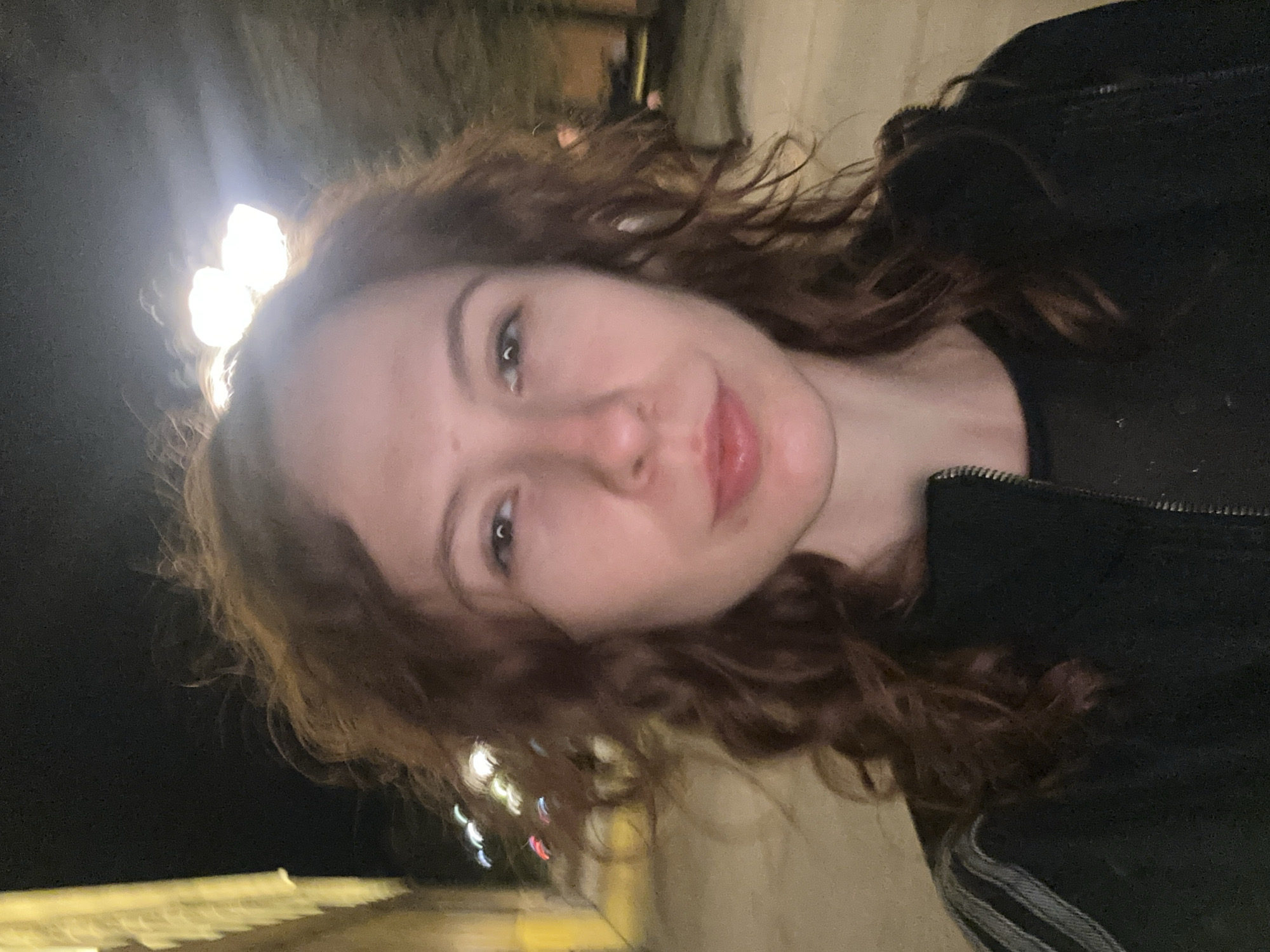 A close-up selfie portrait of a young woman with auburn curly hair, fair skin, and green eyes. She has a slight smile. She is wearing a black jacket. The background is dark, likely night. Bright lights are visible blurred in the upper left corner, suggesting street lamps or city lights. The location is suspected to be Novi Sad, Serbia. The woman is likely standing outside at night in an urban environment.