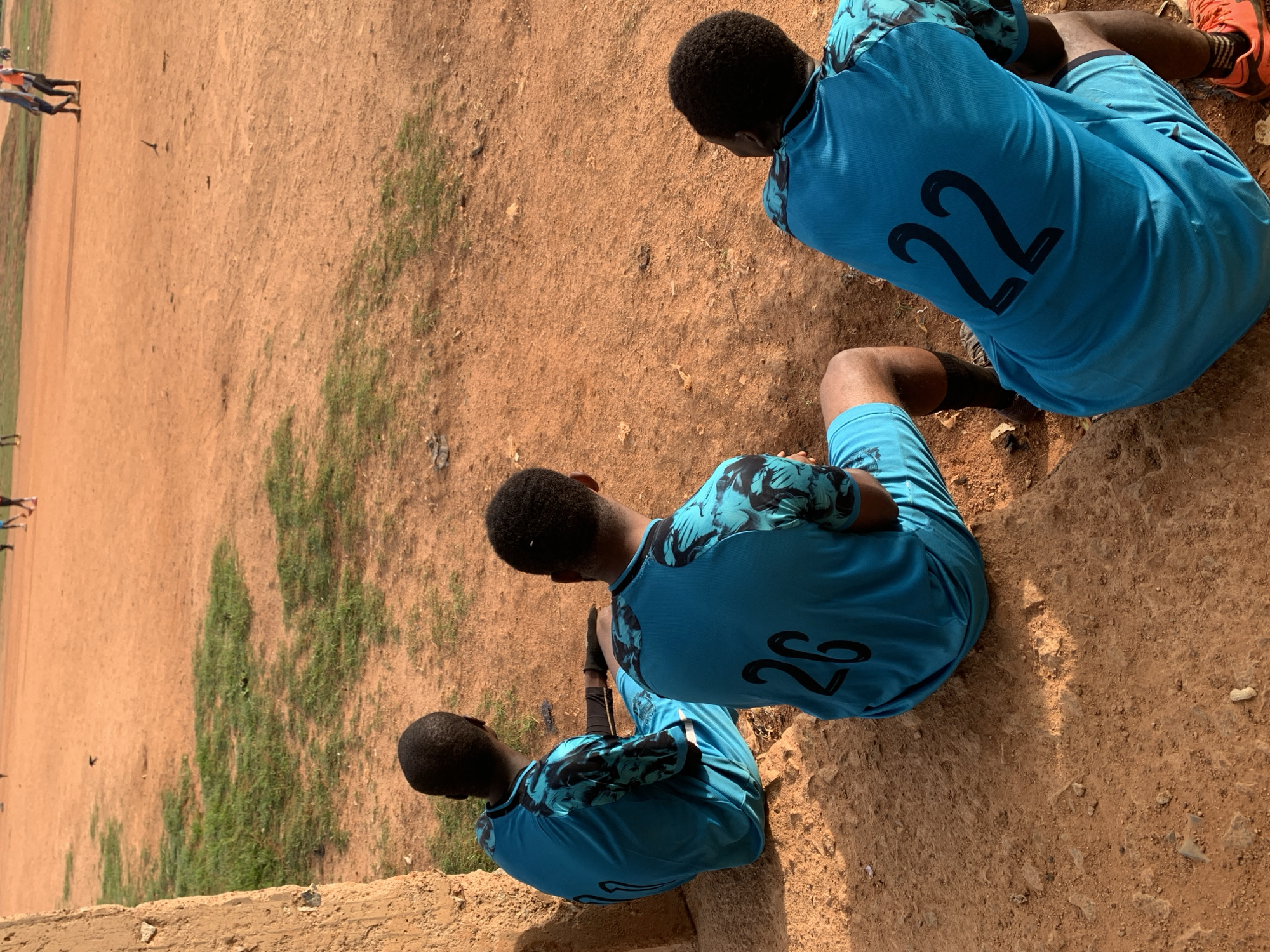 In Bukuru, Nigeria, three young men, likely soccer players, are seated on a dirt ground next to a rough, earthy structure. They are all wearing matching turquoise jerseys with black floral patterns on the sleeves and shorts, and black numbers on their backs. The man on the left wears number 4, the middle man wears number 26, and the man on the right wears number 22. The ground is reddish-brown dirt with patches of sparse green grass. In the background, other figures are visible on a soccer field, suggesting an ongoing game or practice. The overall scene depicts a moment of rest during a sporting activity in an outdoor environment.
