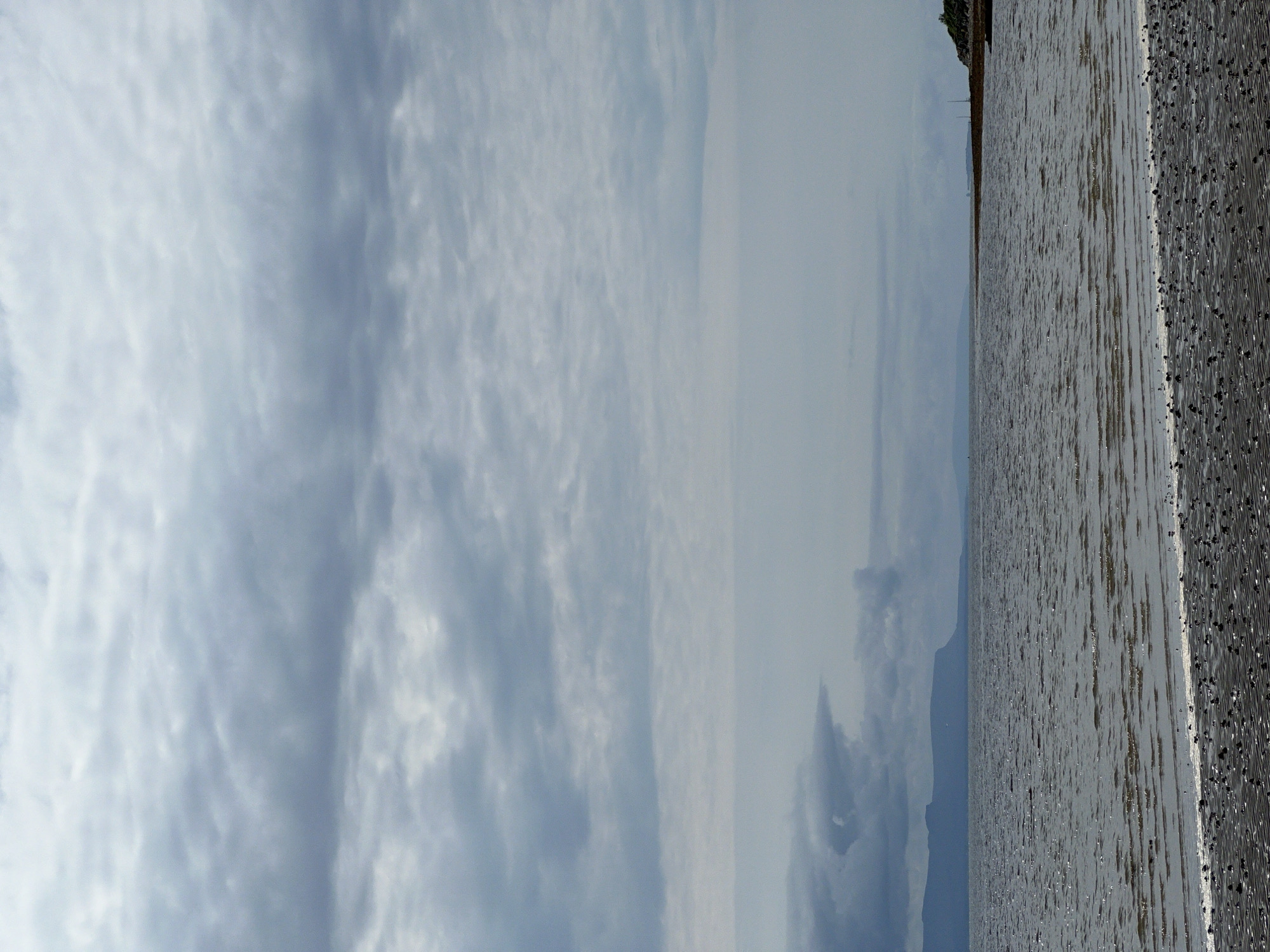 The image depicts a serene coastal landscape on a cloudy day. In the foreground, a textured beach with visible pebbles and sand stretches towards the water. The sea, appearing dark and slightly choppy, occupies the middle ground, reflecting the overcast sky. In the distance, a faint outline of mountains can be seen on the horizon, adding depth to the scene. The sky is dominated by layers of grey and white clouds, suggesting a temperate climate. The overall impression is one of natural beauty and tranquility, with no human presence or activity evident in the frame. The scene is likely located in a rural coastal area, possibly near Mountcharles, Ireland, given the typical geography and weather patterns of the region.