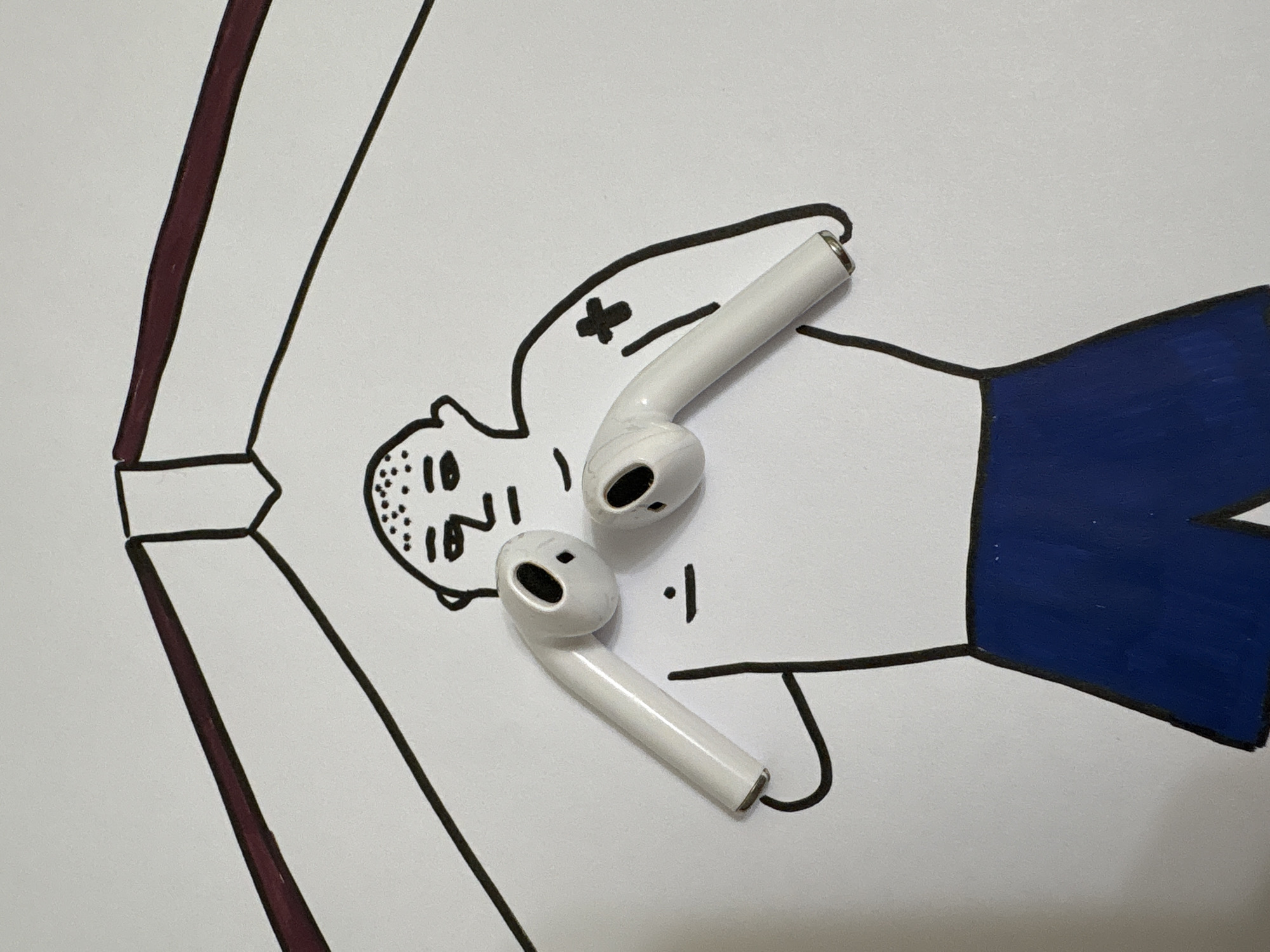 The image depicts a hand-drawn cartoon figure on a white surface, presumably paper. The figure is lying down, drawn with simple black outlines. The head is to the left and features a sparse pattern of dots, possibly representing hair or a skin condition. A cross and a short line are drawn above the figure's chest. The figure is wearing blue shorts. Two white wireless earbuds are placed on top of the drawing. There is no indication of the city of Chaikovsky, Russia, or any other specific location. The scene is sterile and appears to be staged for the photograph.