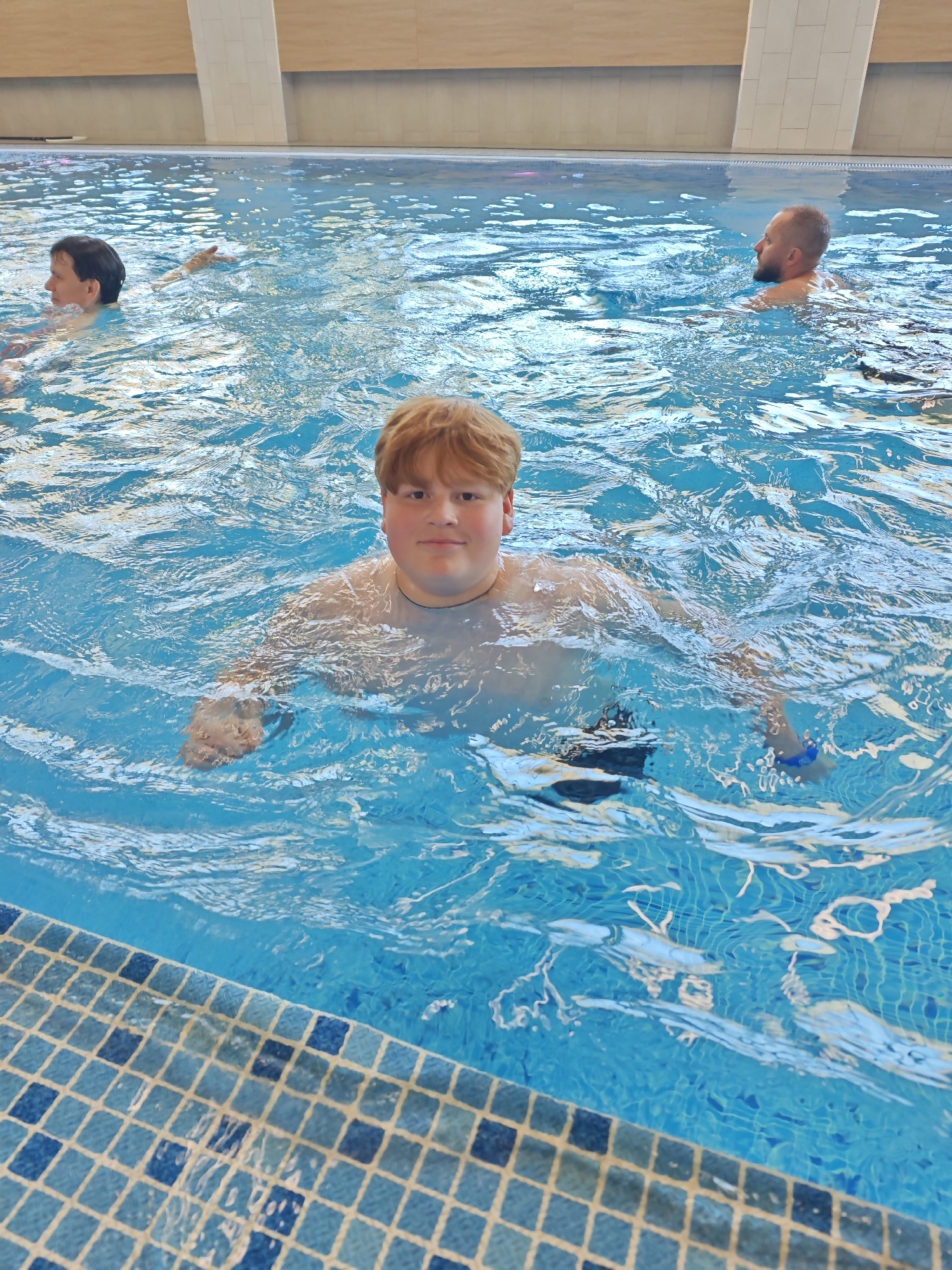 The image shows an indoor swimming pool. A young man with red hair is in the foreground, chest-deep in the water, looking directly at the camera. Two other men are visible in the background, also in the water. The pool is tiled with blue and white mosaic tiles along the edge. The pool is located in a building with a light-colored wall and ceiling. This image was taken in the city of Votkinsk, Russia.