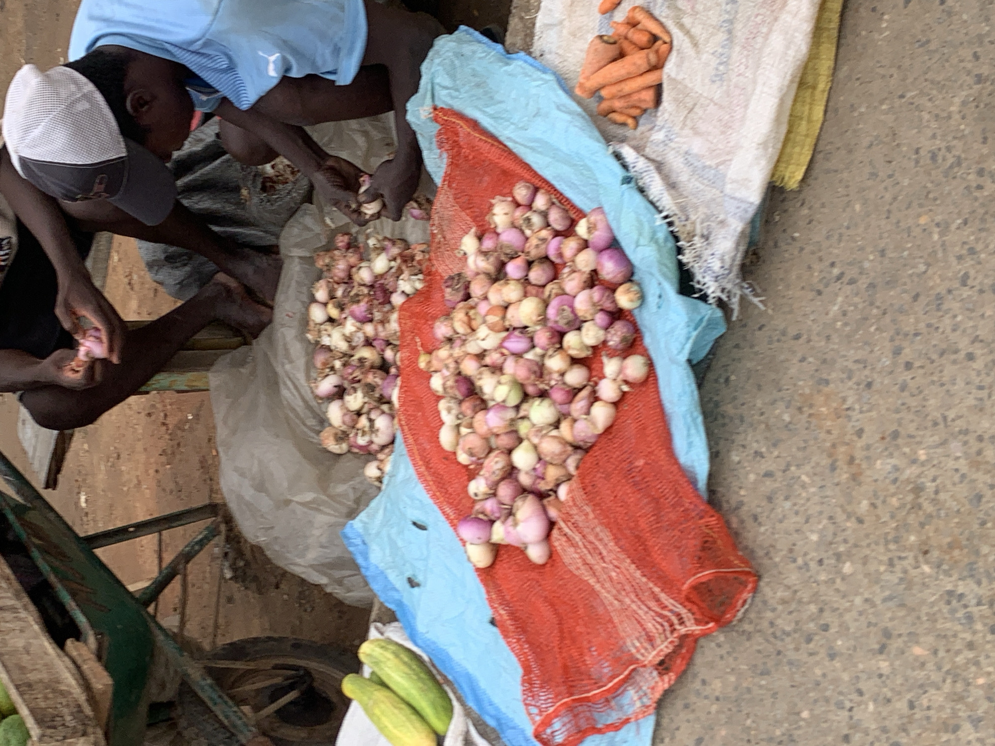 Two individuals are seated on the ground in an open space that appears to be a street or market area in Ikorodu, Nigeria. The persons are handling what seem to be small onions. The onions are in groups on the ground and in an orange mesh bag laid upon a light blue sheet. Additional goods for sale, likely including carrots and cucumbers, are nearby. The ground is concrete. A section of a green metal cart is visible to the lower left.