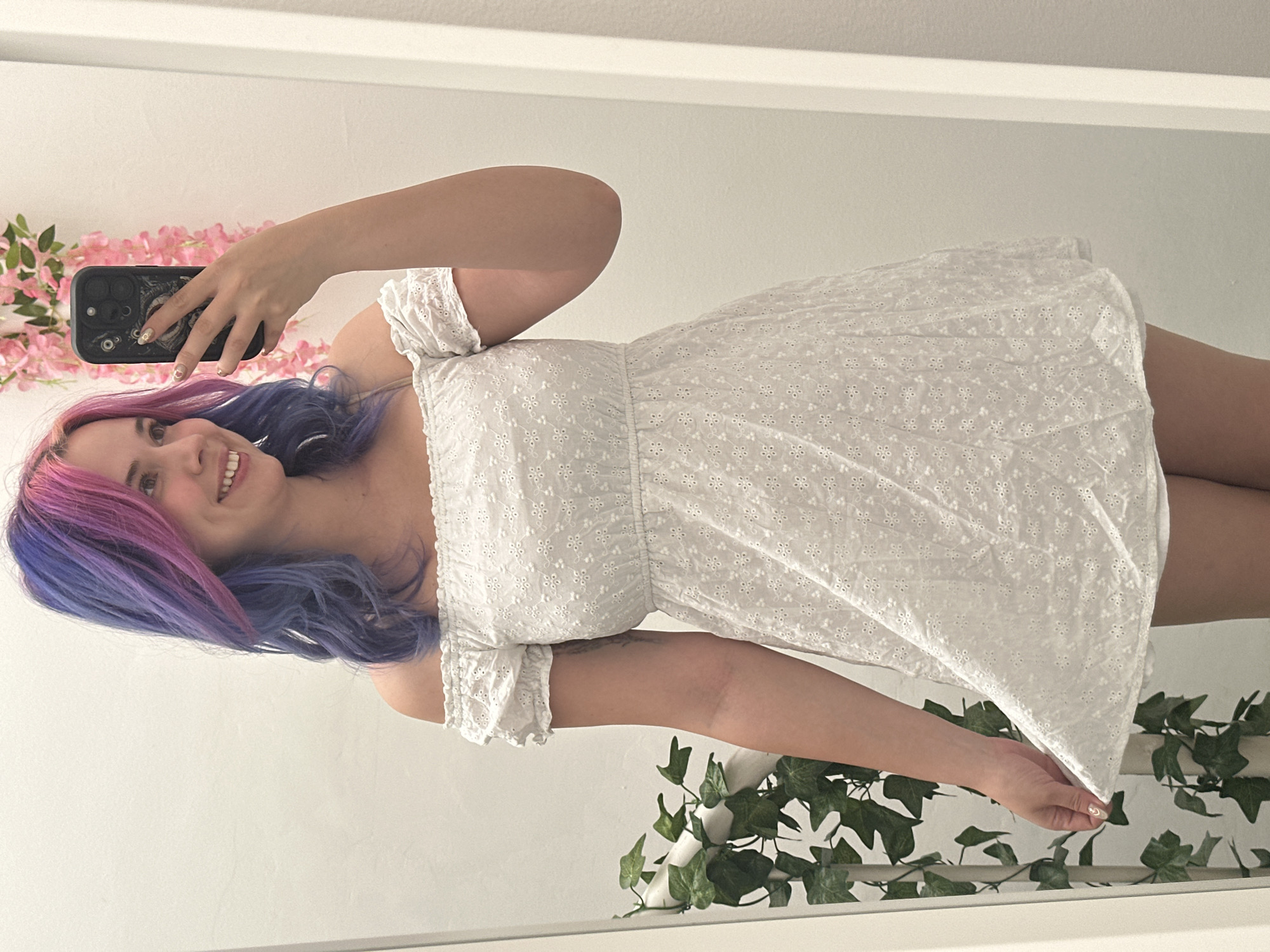 A young woman with a cheerful expression and a wide smile is captured in a mirror selfie, showcasing her unique shoulder-length hair dyed in vibrant pink and purple/blue hues. She is wearing a white off-the-shoulder sundress adorned with intricate eyelet embroidery, featuring small floral or geometric patterns. In her right hand, she holds a smartphone with a dark case that appears to have a distinctive character or monster design, positioned to take the photo. Her fingernails are manicured with a light, subtle design.
The setting, reflected in the mirror, reveals a clean, plain white wall in what appears to be an indoor space in the City of Zagreb, Croatia. Decorative elements are visible: a string of artificial pink flowers is draped to the left, and artificial green ivy is arranged along the bottom right, suggesting a shelf or ledge. The lighting is bright and even, indicating either natural daylight or well-diffused artificial light. There is no visible text in the image.