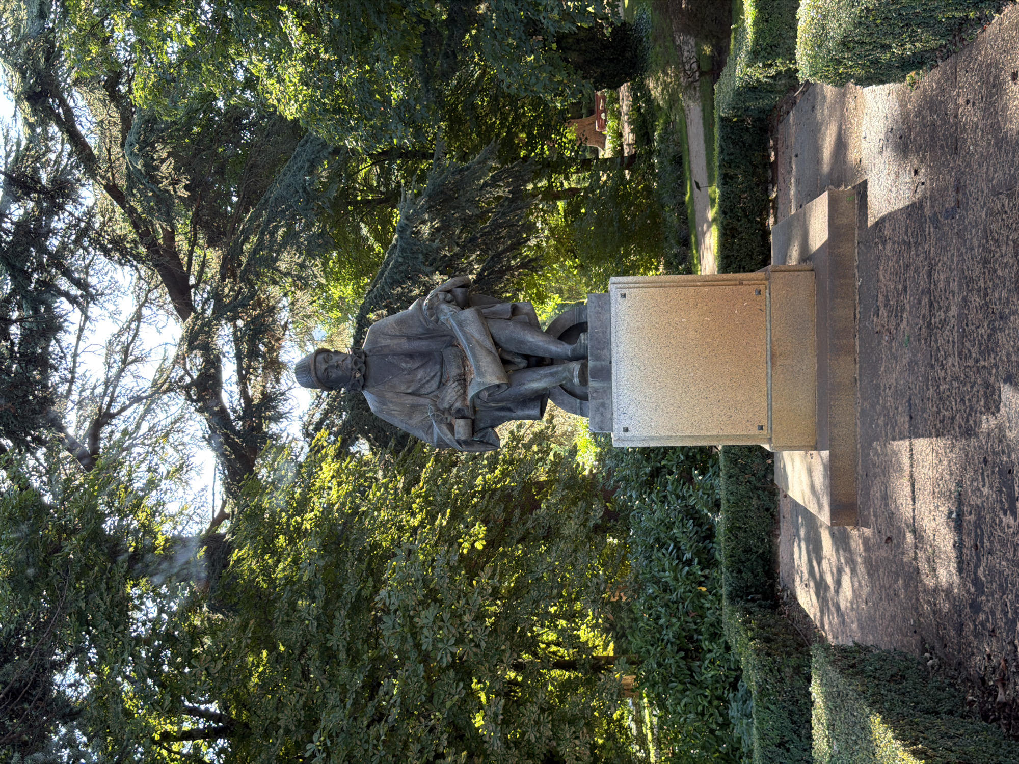 The image shows a bronze statue of a man seated on a stone pedestal, located outdoors in a park setting. The man is wearing historical attire, including a hat and a ruffled collar, and is holding a scroll in his hands. The statue is situated in the Royal Site of San Lorenzo de El Escorial, Spain, specifically within the Jardines de la Casita del Infante. The surrounding environment consists of lush green trees, manicured hedges, and a paved walkway bathed in sunlight. The sunlight creates distinct shadows on the ground, indicating it is daytime. There are no other people visible in the immediate vicinity of the statue. The overall scene is peaceful and serene, highlighting the statue as the central subject amidst the natural landscape.