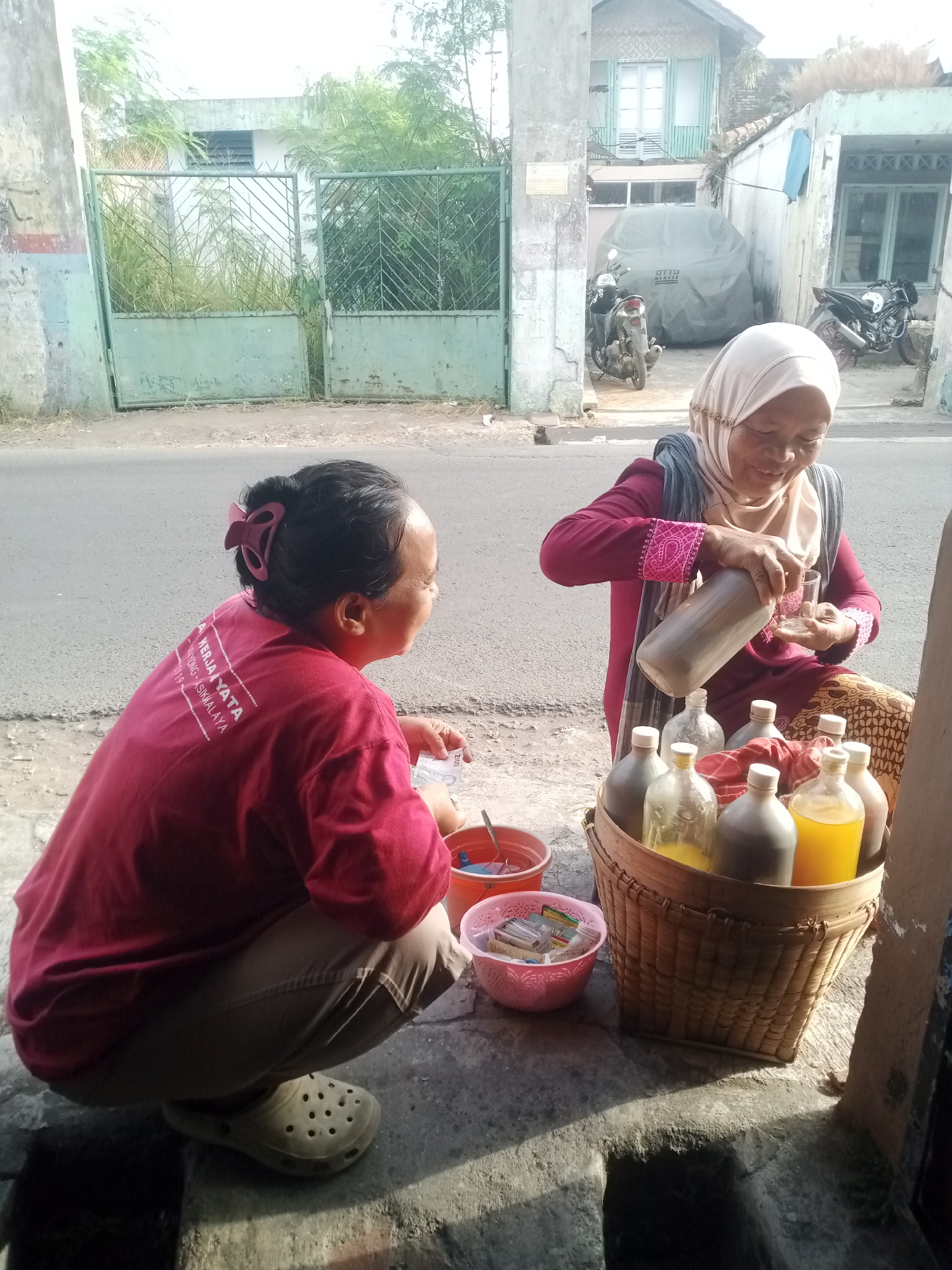 Two Indonesian women are crouched down on a street in Tasikmalaya, Indonesia, near a building with a green gate and a light-colored facade. The woman on the right, wearing a maroon shirt and a light-colored headscarf, is pouring a brown liquid from a large glass bottle into a small glass. In front of her is a woven basket filled with several glass bottles containing various colored liquids, some yellow and some dark brown. A red cloth is draped over some of the bottles. To her left, a woman wearing a red t-shirt and light brown pants is holding a small stack of paper money and appears to be interacting with her. In front of her is a pink bowl containing small packets. The street appears to be paved, and a sidewalk is visible. In the background, there are motorcycles parked near the building. The scene suggests a street-side vending or exchange taking place.