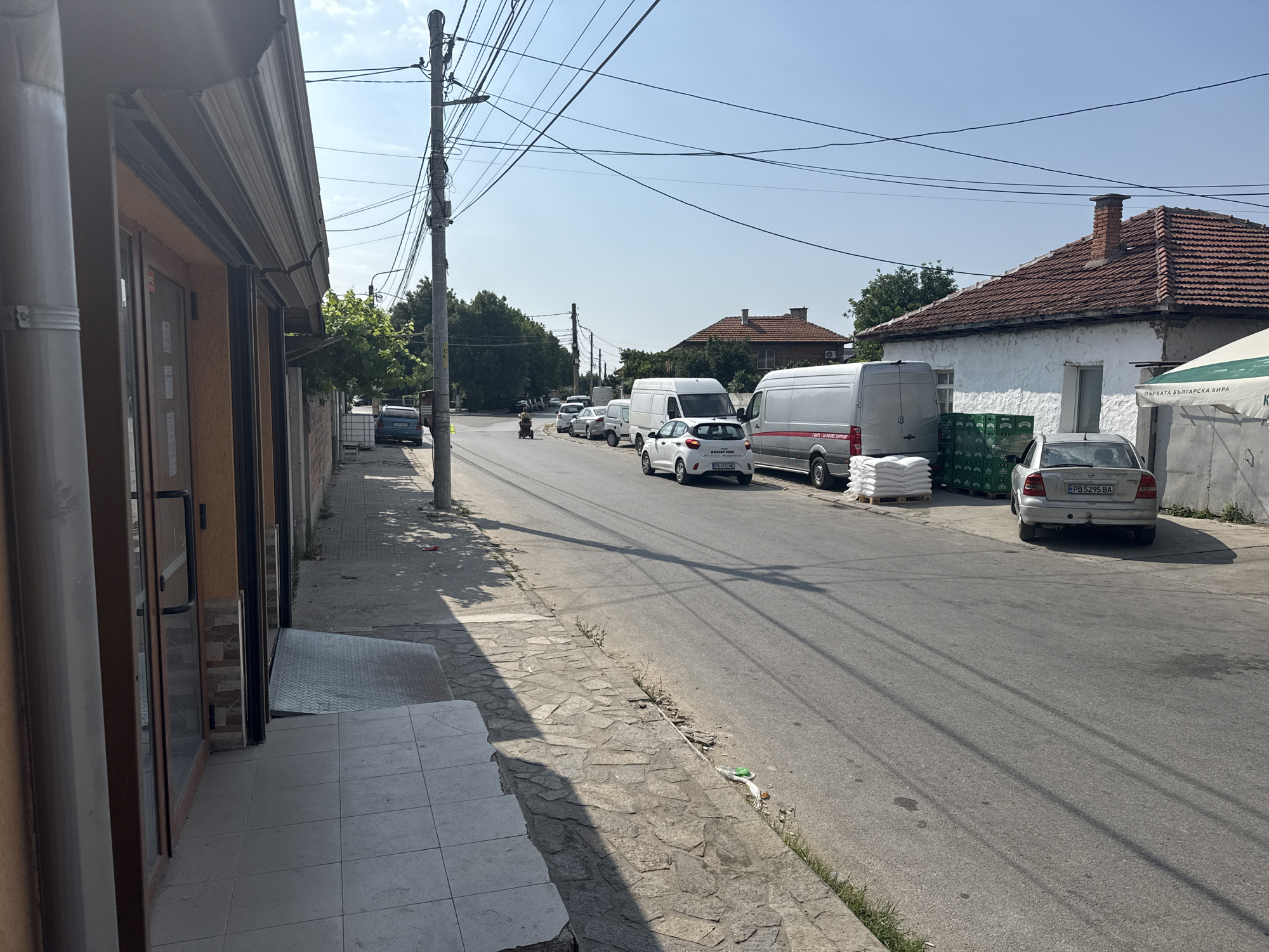 The image depicts a street scene in Rakovski, Bulgaria. On the left, the side of a building with an orange facade and glass doors is visible, with a covered awning and drainpipe. A tiled pathway leads away from the building. The street is paved with asphalt, and a sidewalk made of irregular stone slabs runs alongside it. Several vehicles are parked or driving on the street, including a silver car, two white vans, and other cars. Stacks of white bags and green crates are piled on pallets on the side of the road. Overhead, numerous electrical wires crisscross the sky, supported by a tall utility pole. The background features residential buildings with tiled roofs and lush green trees. The sky is clear and blue, indicating a sunny day.