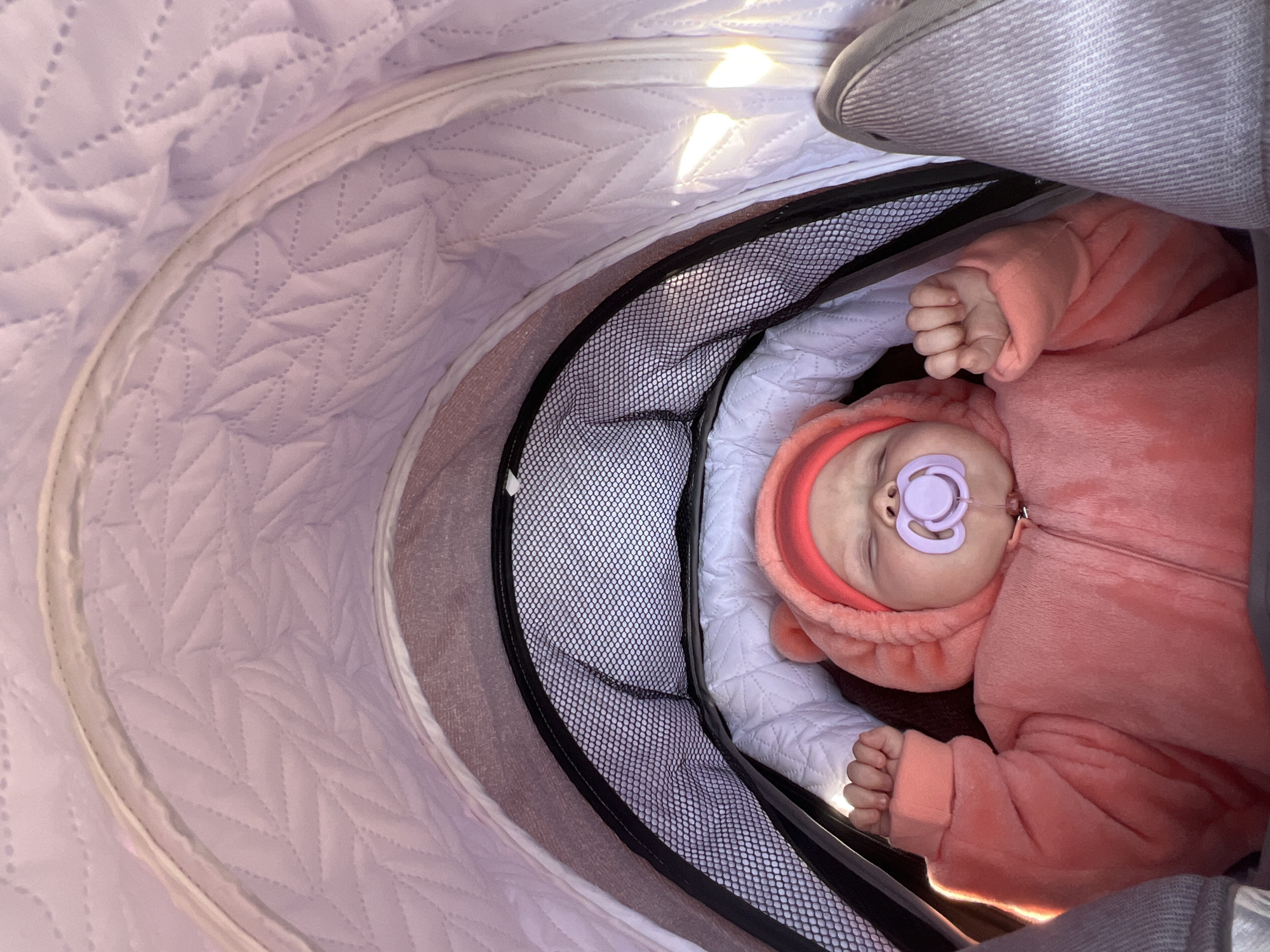 A baby sleeps in a stroller. The baby wears a pink jumpsuit with a hood. The stroller has a quilted lilac interior and a black mesh sunshade. The location is unknown but could be Votkinsk, Russia.