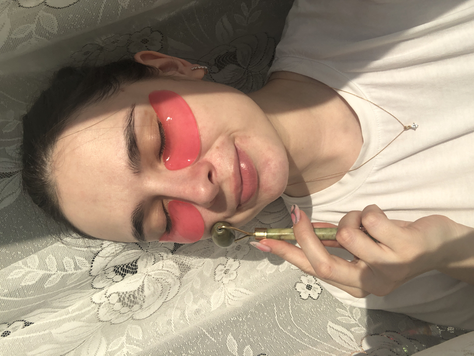 A young woman is depicted relaxing indoors in Noviy, Russia, lying down on a surface covered with a white, intricate lace-patterned fabric, possibly a bed or sofa. She is engaged in a skincare routine, with her eyes closed, and two glossy, pink hydrogel eye patches positioned under her eyes. Her right hand holds a green jade facial roller, which she is using on her right cheek.
The woman has dark hair and a fair complexion, with a slight, contented smile on her lips. She is wearing a simple white short-sleeved t-shirt. A delicate gold chain necklace with a small, clear gemstone pendant hangs around her neck, and a sparkling stud earring is visible in her left ear. Her fingernails are manicured with a light base and subtle pink and black floral designs.
The scene is illuminated by bright natural light, likely from a window, as evidenced by distinct shadows cast across her face and the lace fabric. The fabric displays elaborate floral motifs, including larger rose patterns and smaller blossoms, creating a decorative background. The image conveys a sense of calm and self-care during the daytime. No visible text is present in the image.